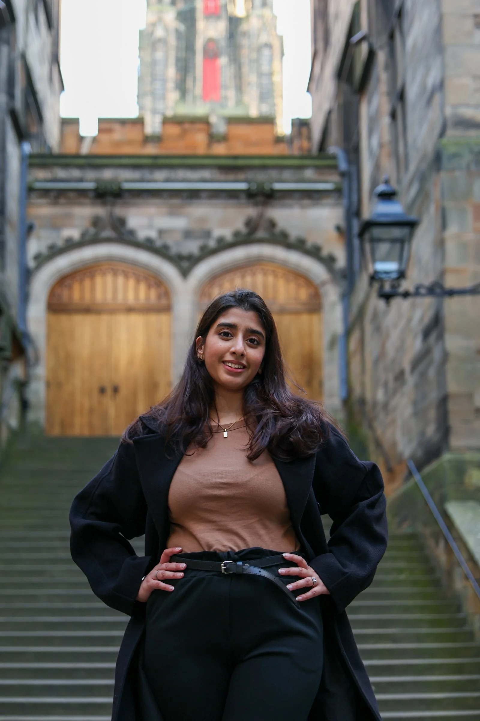 Young woman with dark hair standing on stone steps in urban setting, smiling near stone walls and wooden gates, with a large historic building visible in the background.