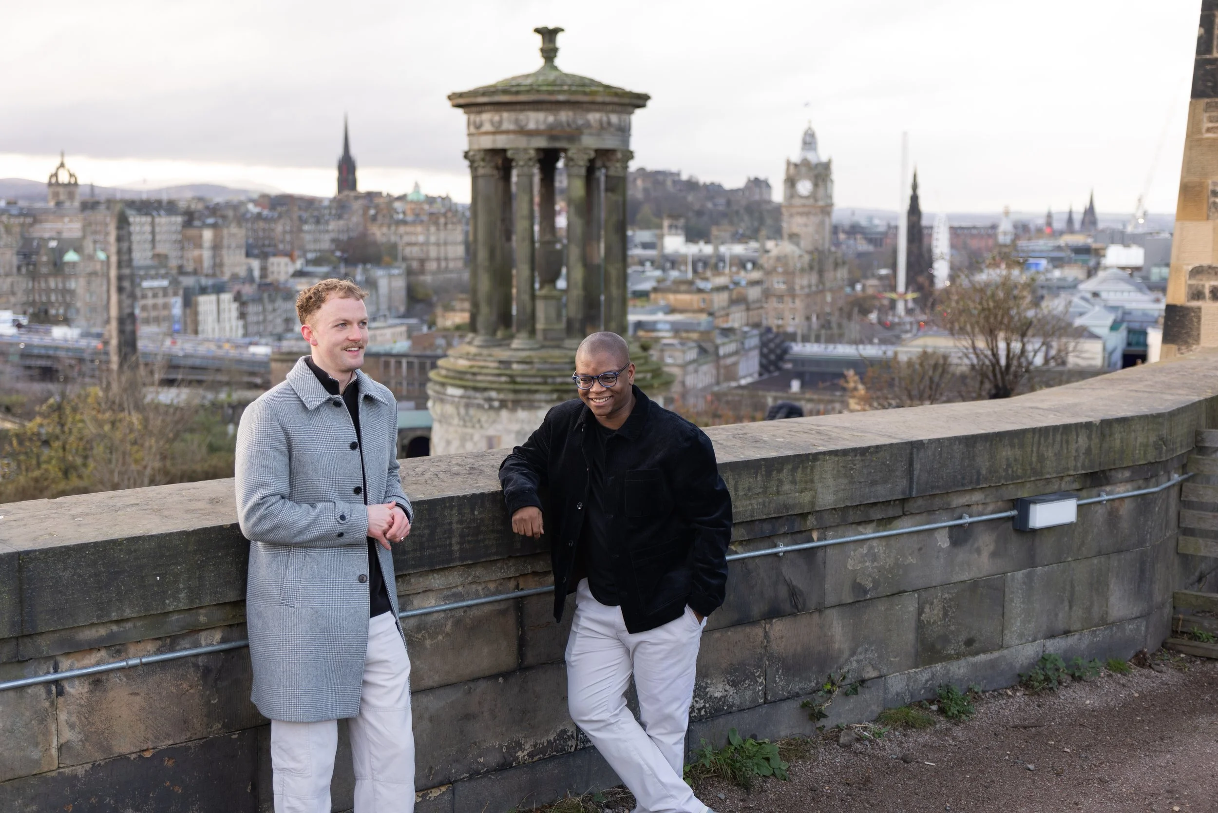 Two men standing outdoors on a stone wall with a cityscape in the background, including historic buildings and a prominent clock tower, during daytime.
