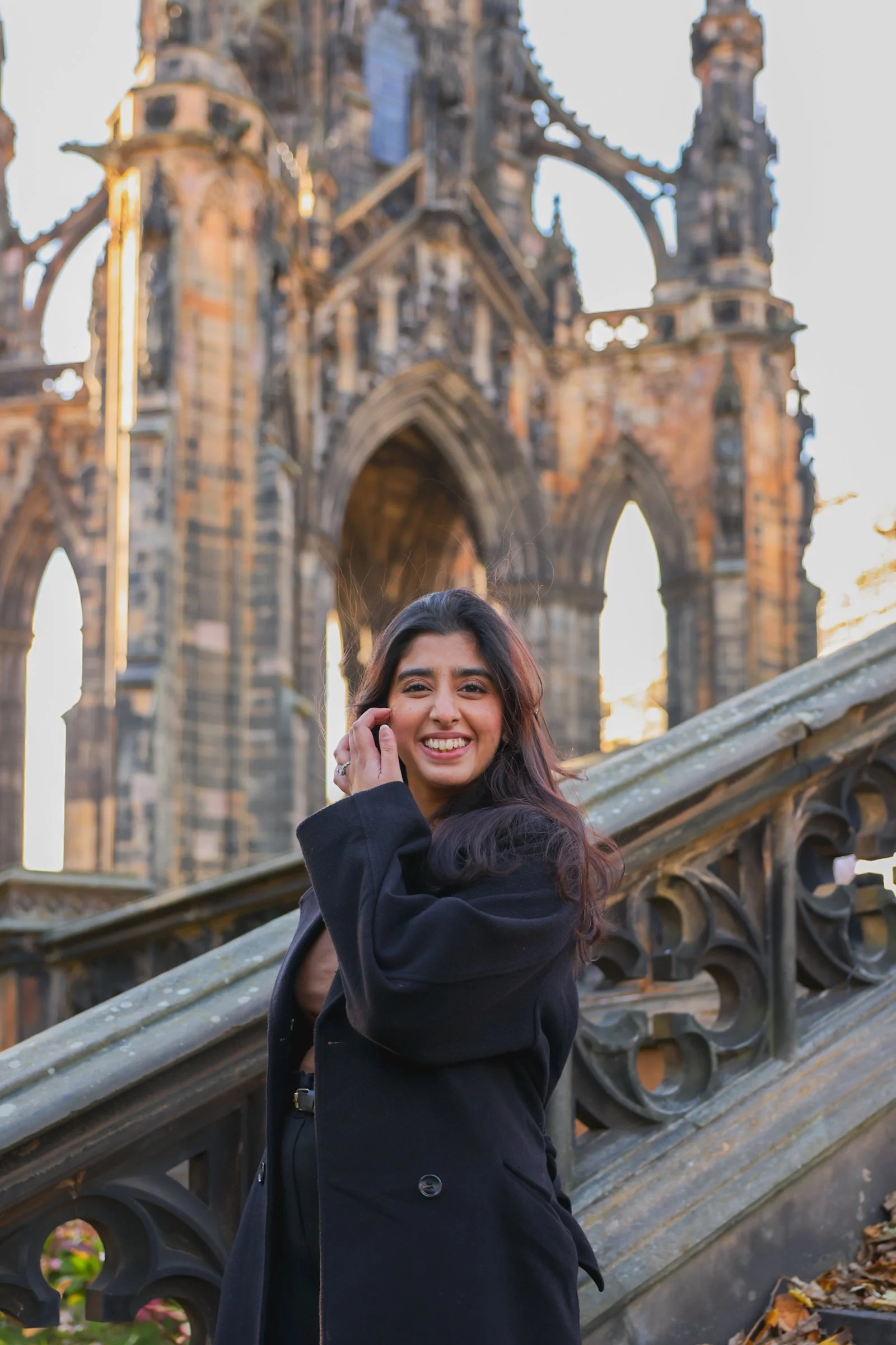 A young woman smiling and talking on her phone, standing on a staircase with a historical Gothic-style church in the background during sunset.