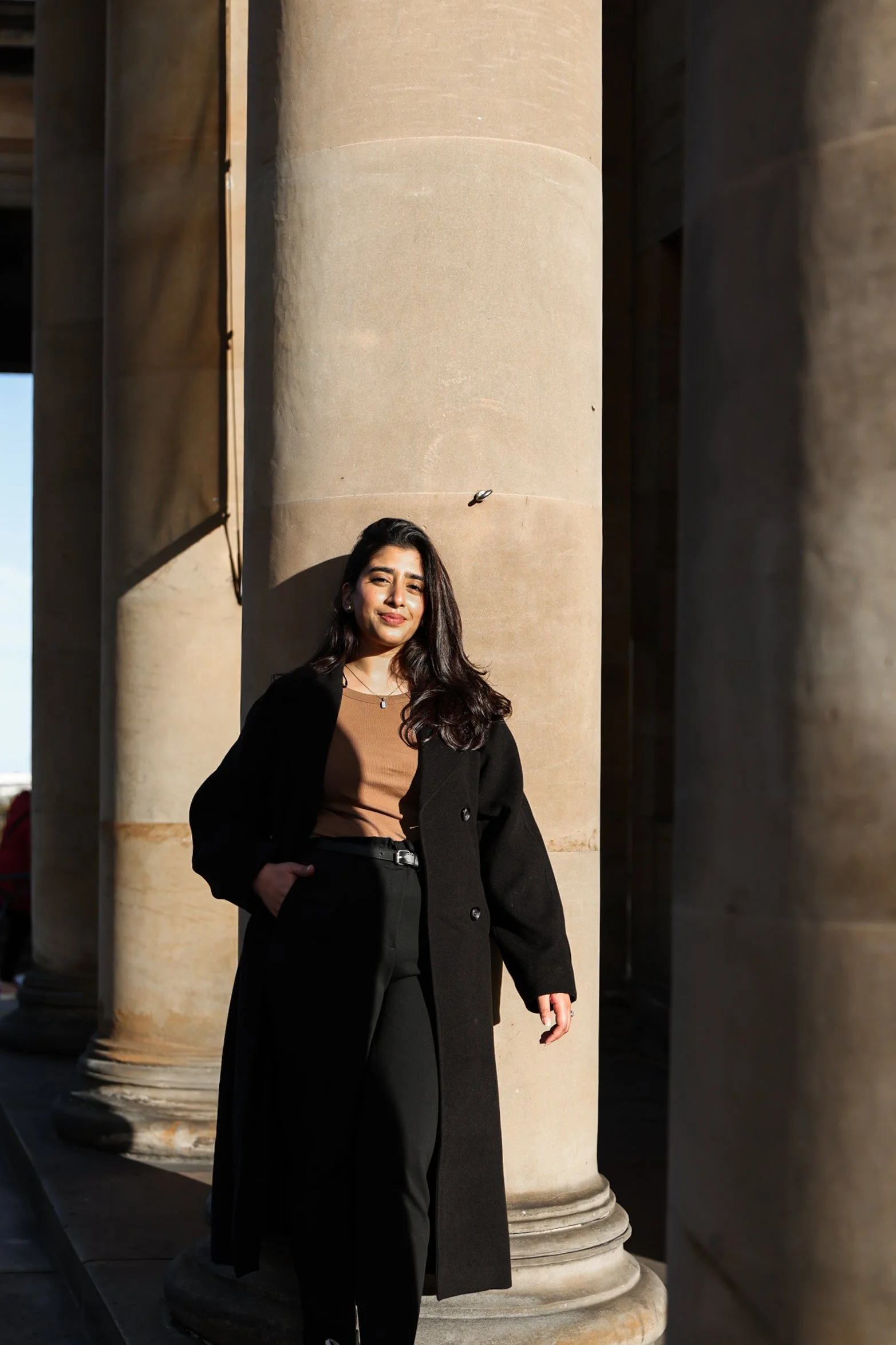 A woman standing next to large stone columns outdoors, dressed in black pants, a brown top, and a black coat, with her hand in her pocket.