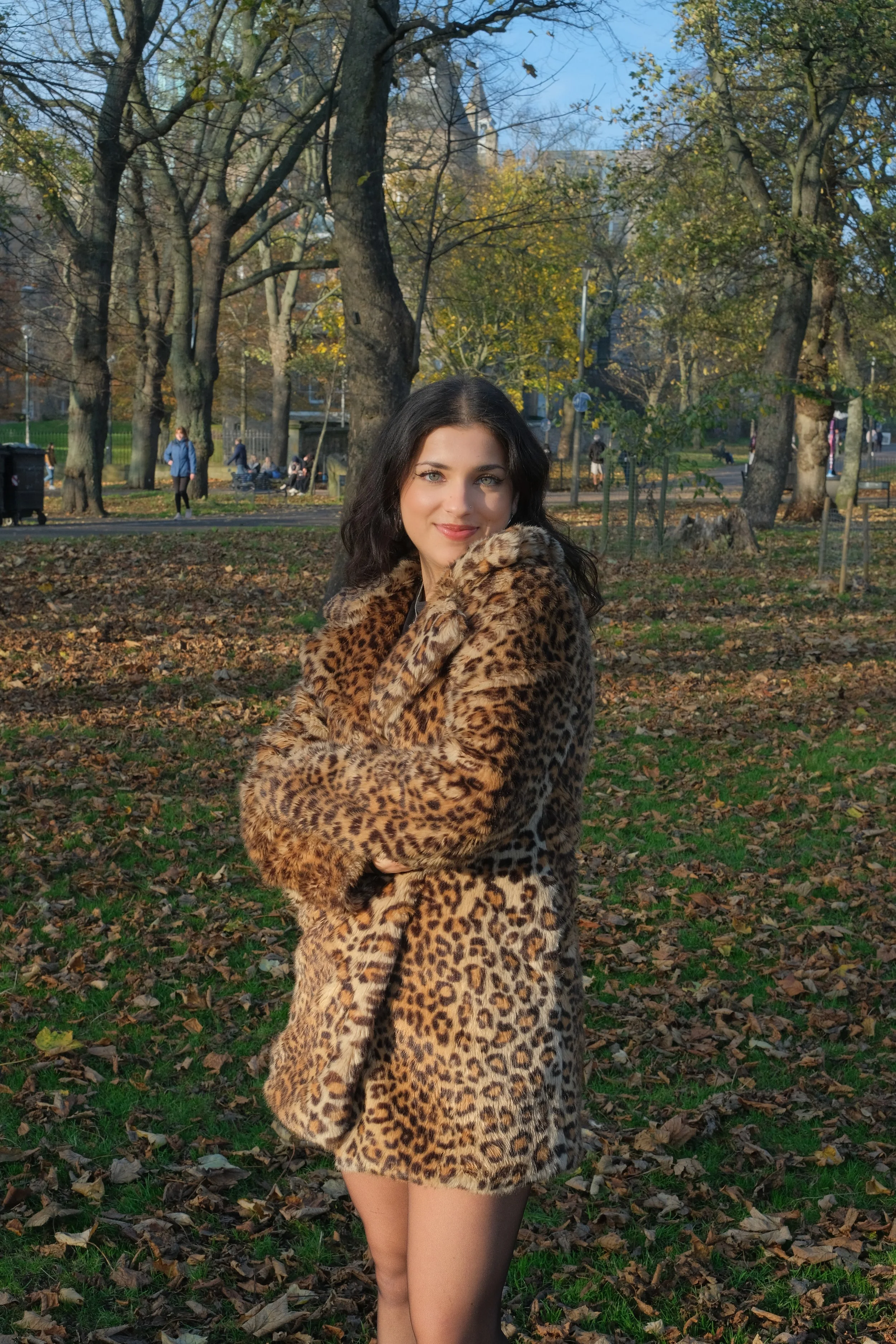 A woman with dark hair smiling while wearing a leopard print coat, standing in a park with trees and fallen leaves during autumn.