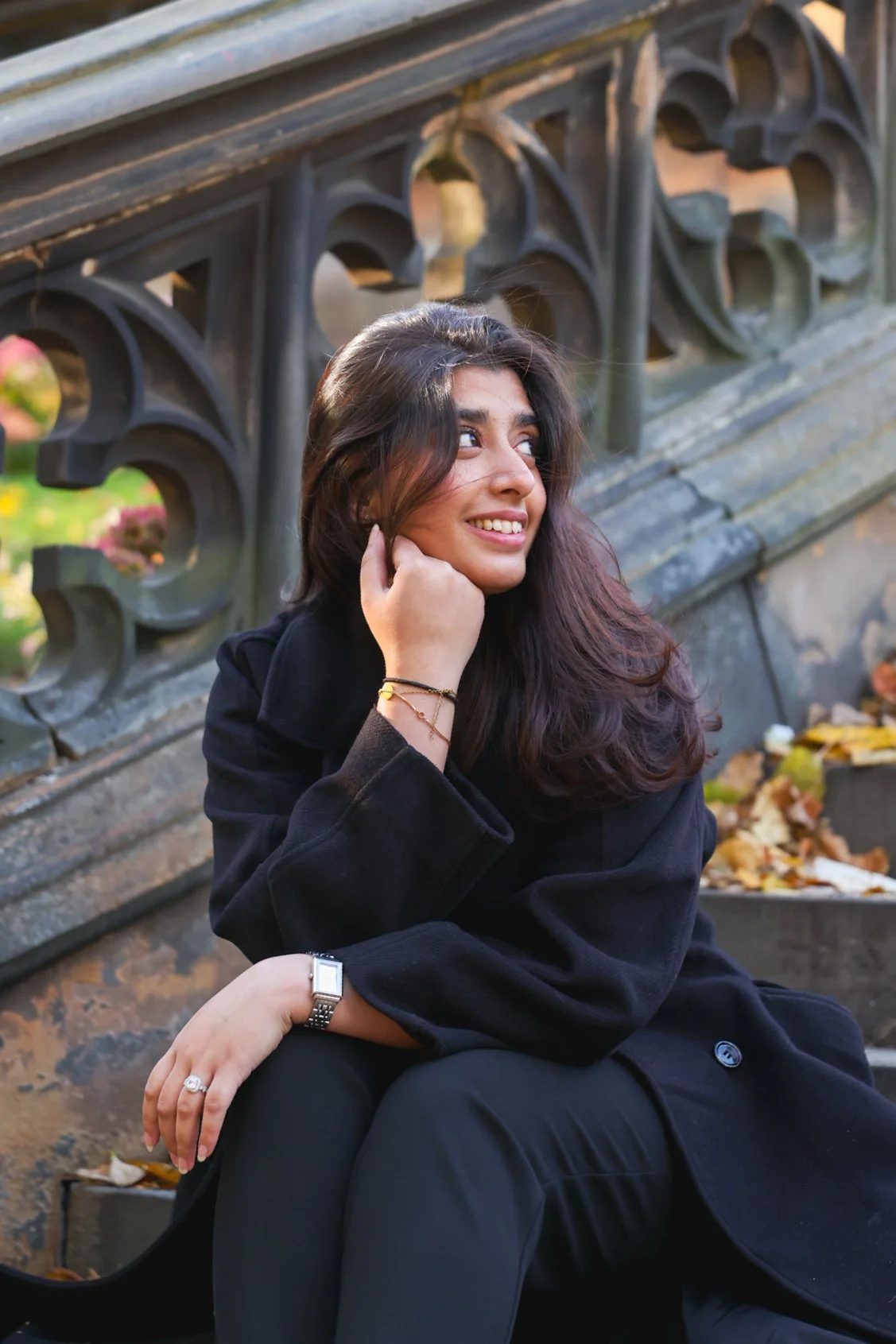 A young woman with dark hair and a black jacket sitting on steps outside, resting her chin on her hand, looking to the side and smiling. There are autumn leaves on the steps behind her and a decorative metal railing.