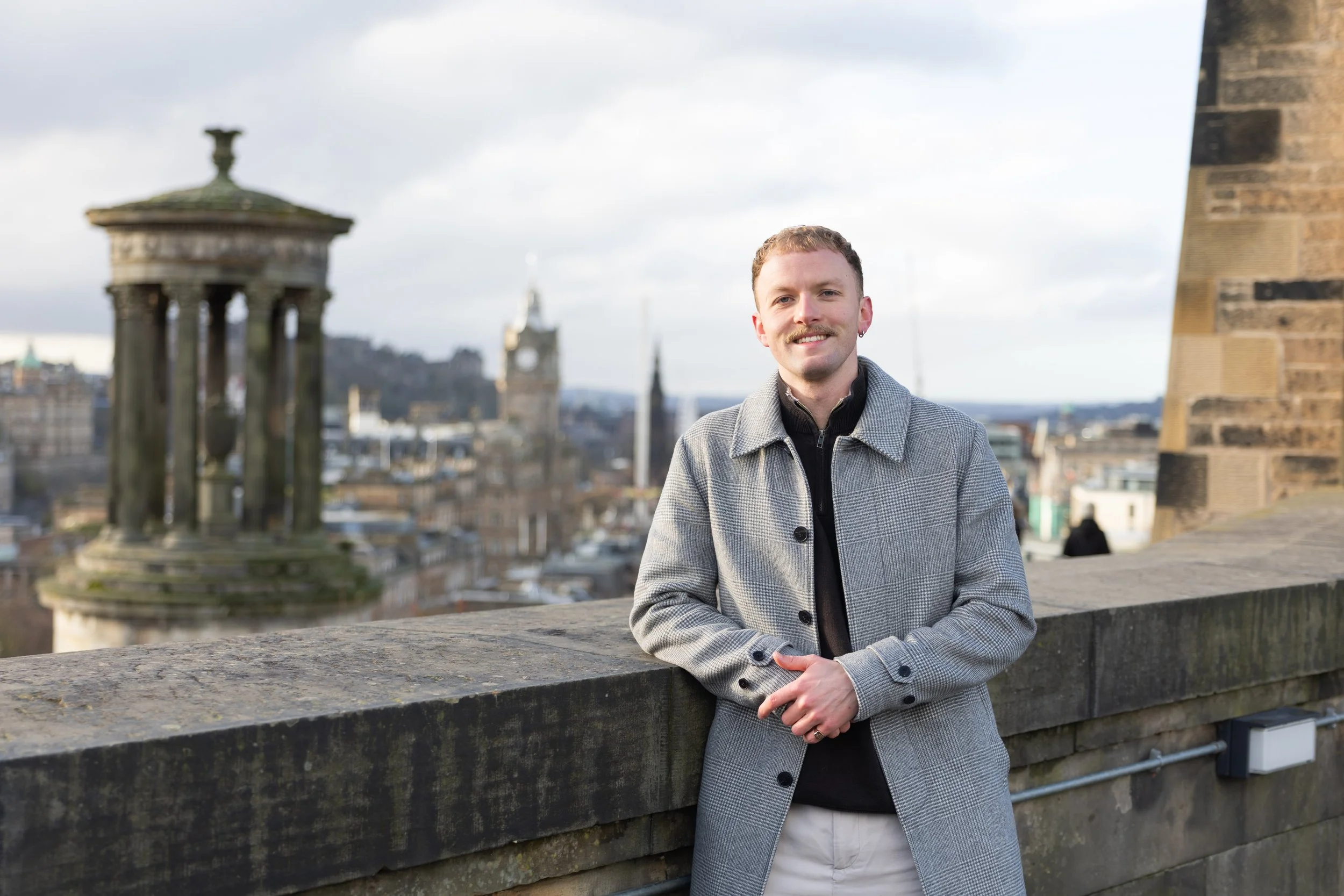 A man with short light brown hair and a mustache smiling while leaning on a stone wall in a city setting. In the background, there are historical buildings, including a rounded pagoda-style structure on the left, and a clock tower with a spire. The s