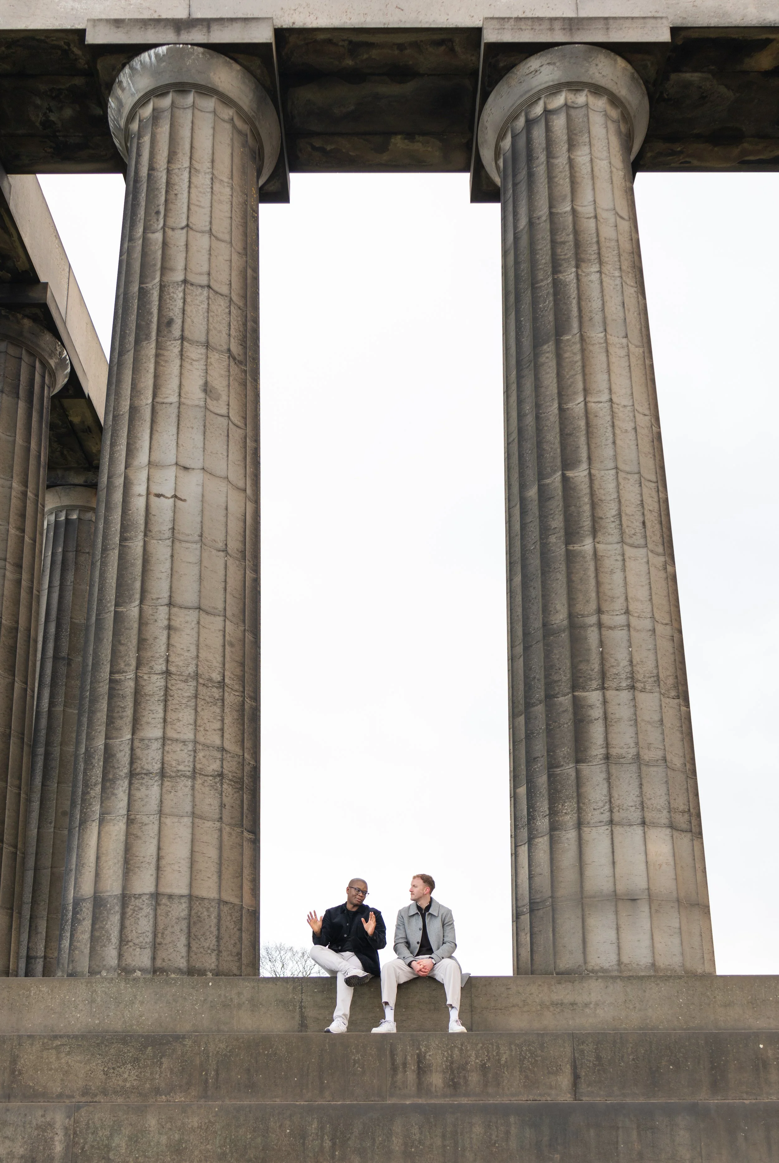 Two men sit on a step under large ancient columns of a historical monument, conversing against a cloudy sky.