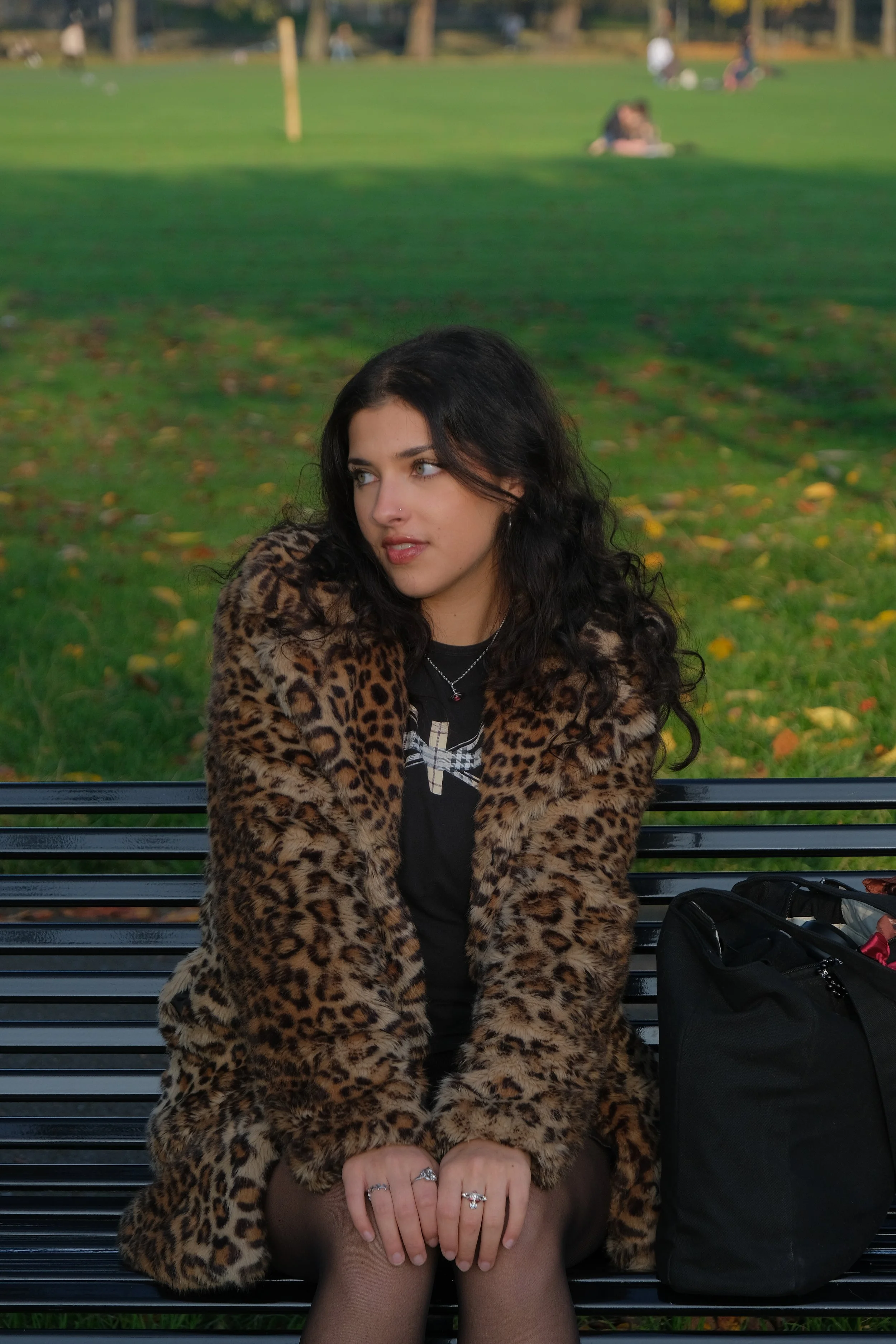 A young woman with black curly hair sitting on a park bench, wearing a leopard print coat, with a black backpack beside her, in a park with green grass and trees in the background.