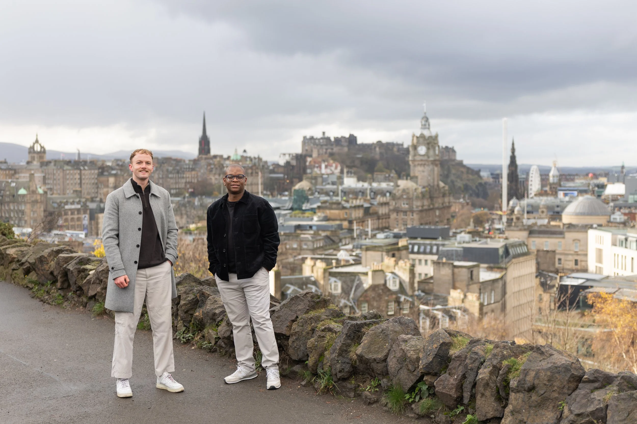 Two men standing on a stone barrier overlooking a cityscape with historic buildings, clock tower, and castle in the background under a cloudy sky.