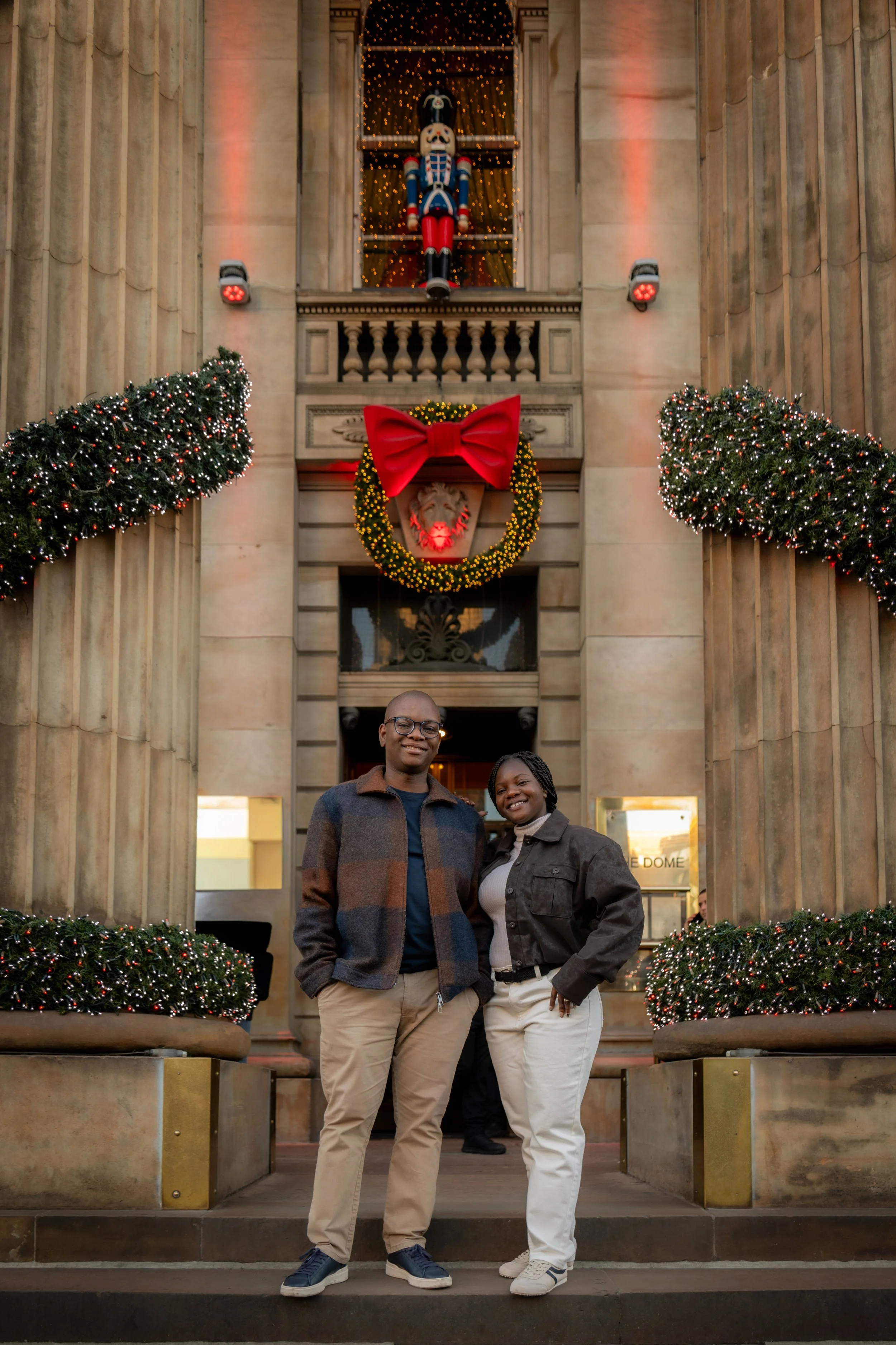 A man and woman standing in front of a decorated building with Christmas ornaments, wreath, and large nutcracker figure, smiling at the camera.