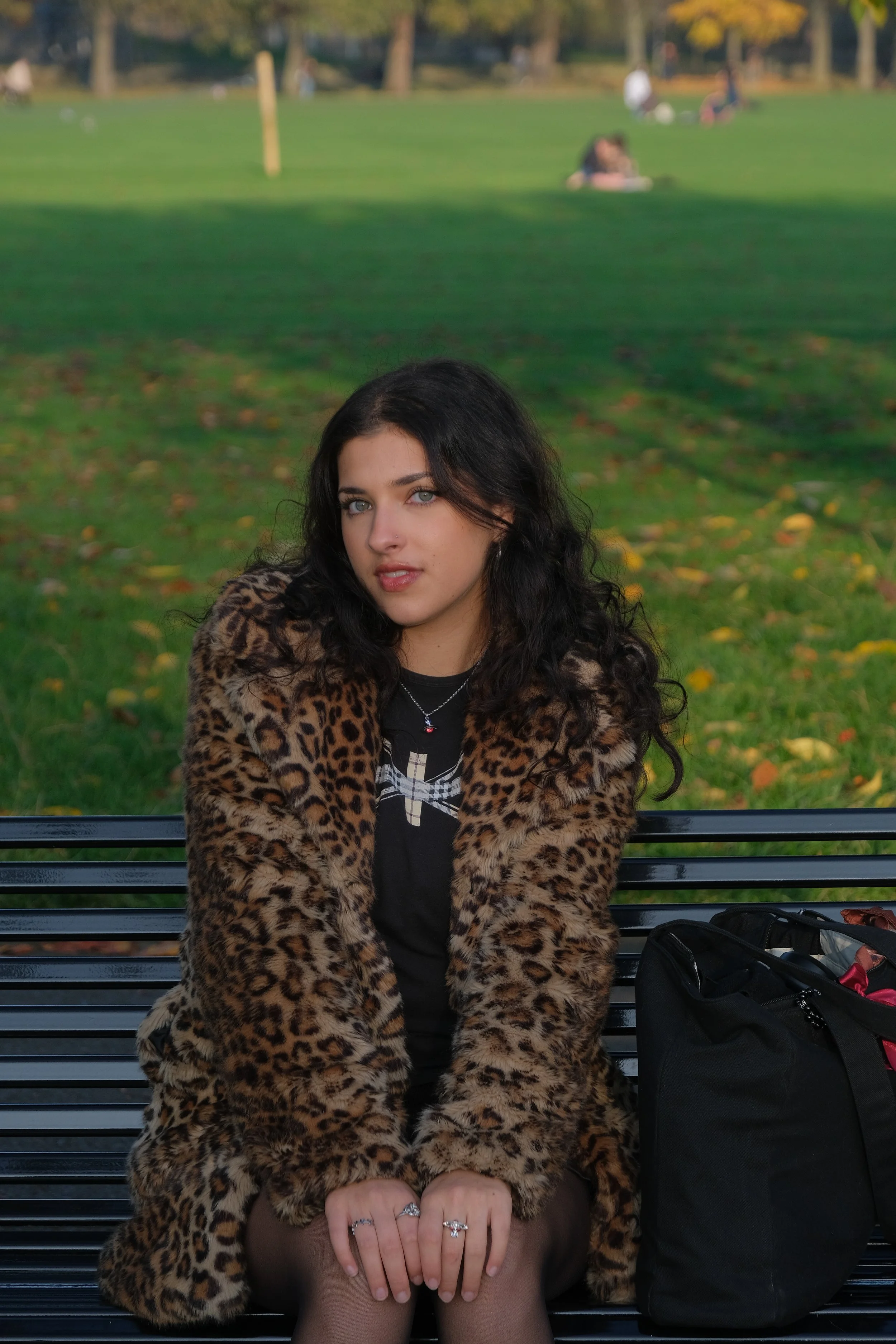 A young woman with dark curly hair and blue eyes sitting on a park bench, wearing a leopard print fur coat, black shirt with graphic design, and jewelry. A black bag is placed beside her on the bench, with a blurred park scene in the background.