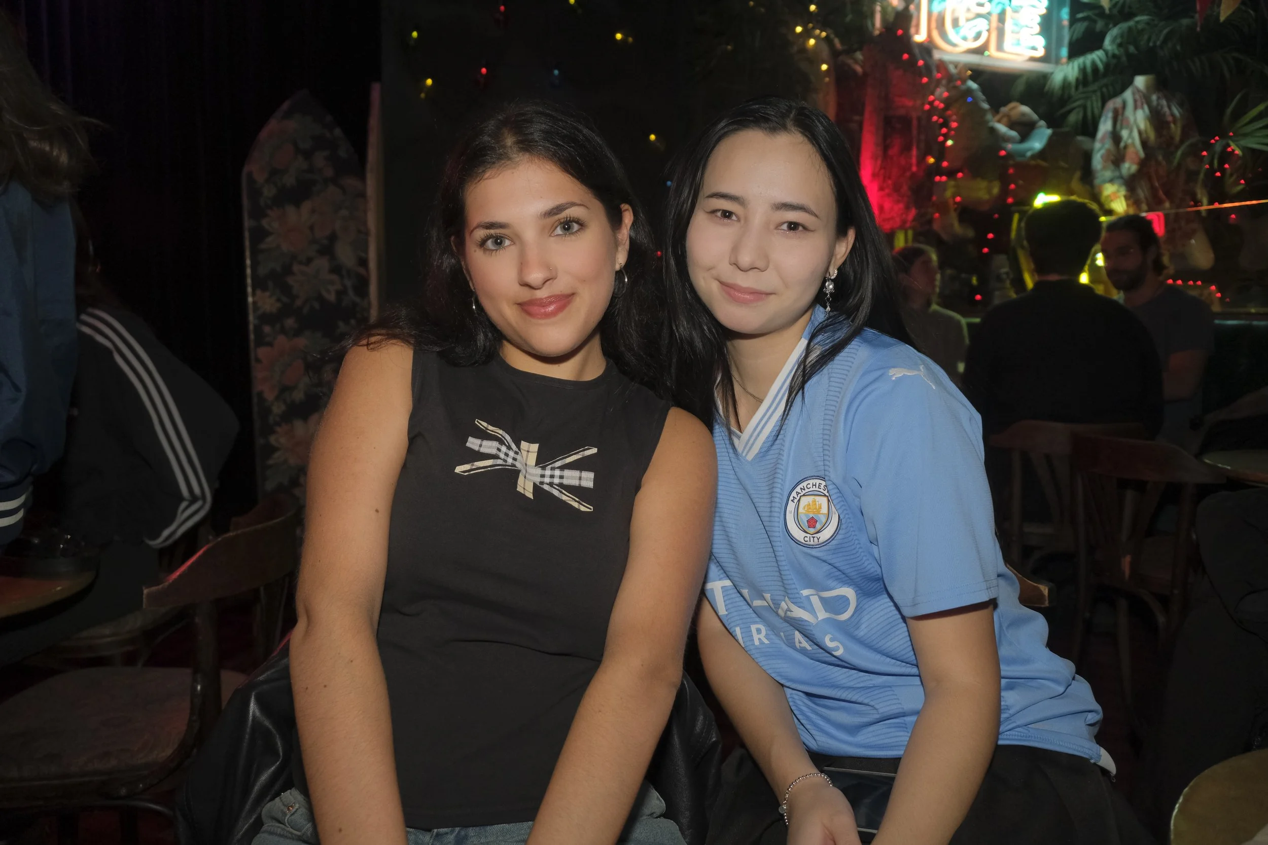 Two young women sitting close together at a dimly lit indoor event or party, smiling at the camera, with colorful lights and blurred people in the background.