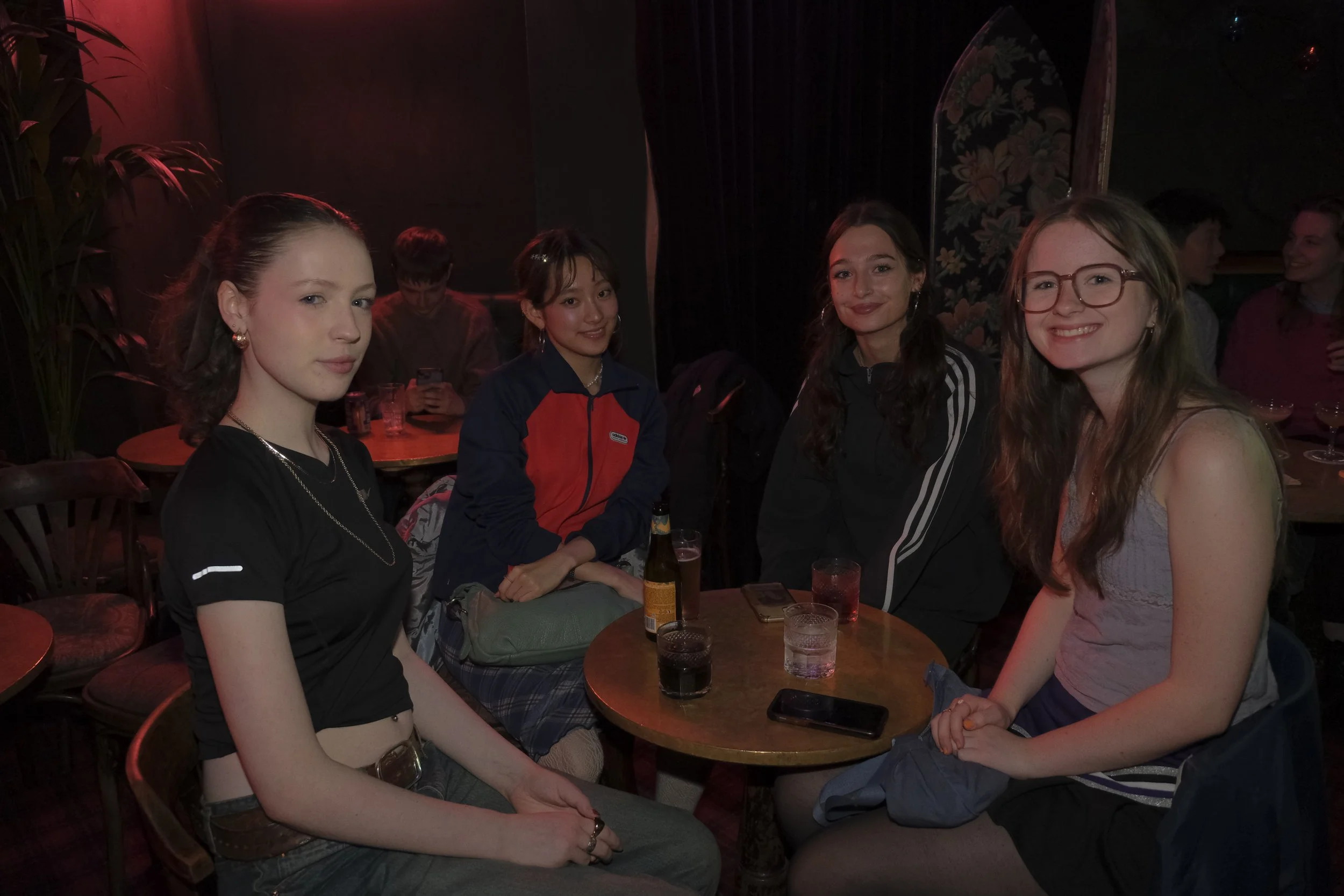 Four young women sitting around a small round table in a dimly lit bar or club, with drinks and mobile phones on the table, smiling and posing for the photo.