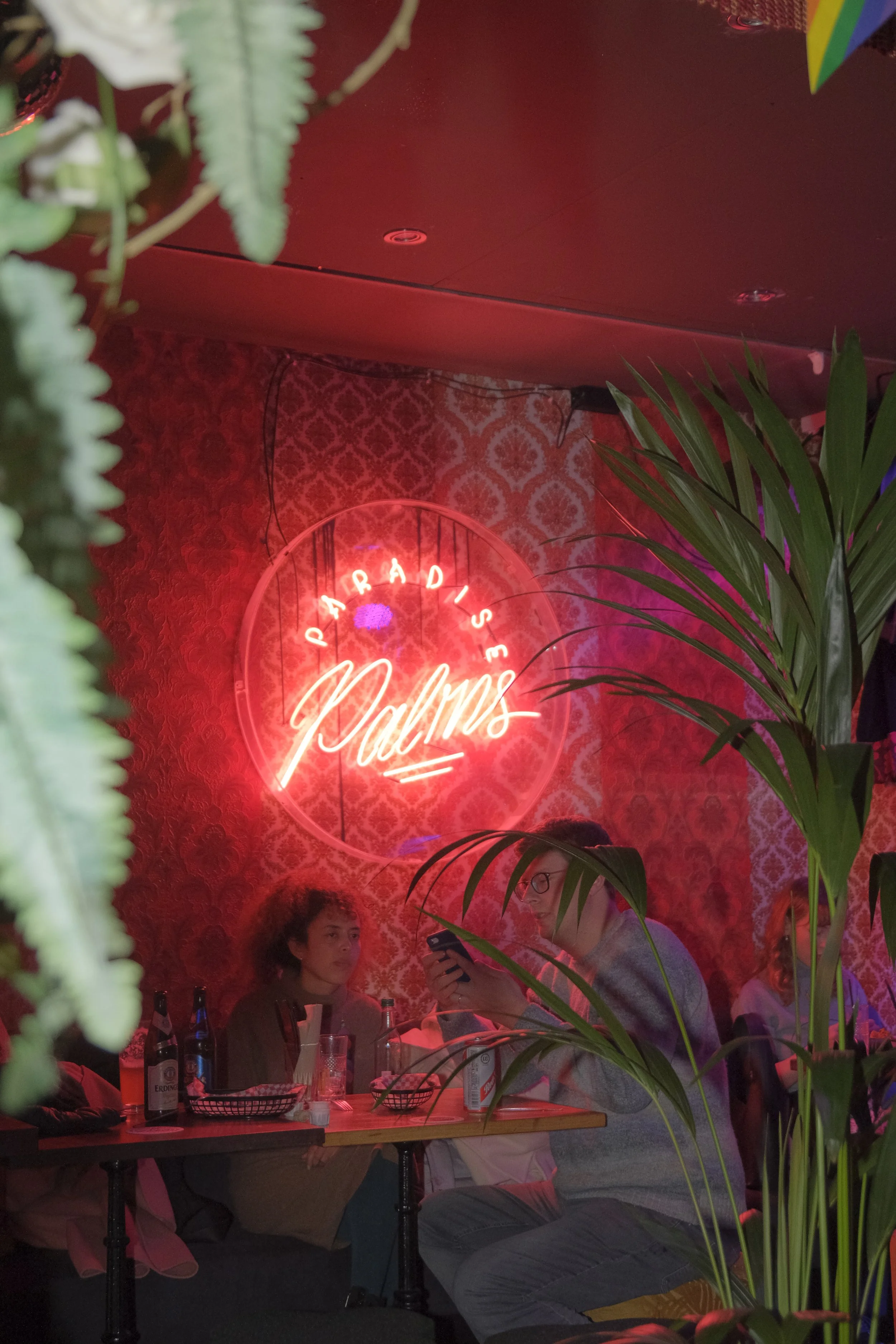 People sitting at a table in a dimly lit restaurant with a neon sign that reads 'Paradis Palm's' on a red patterned wall, decorated with green plants.