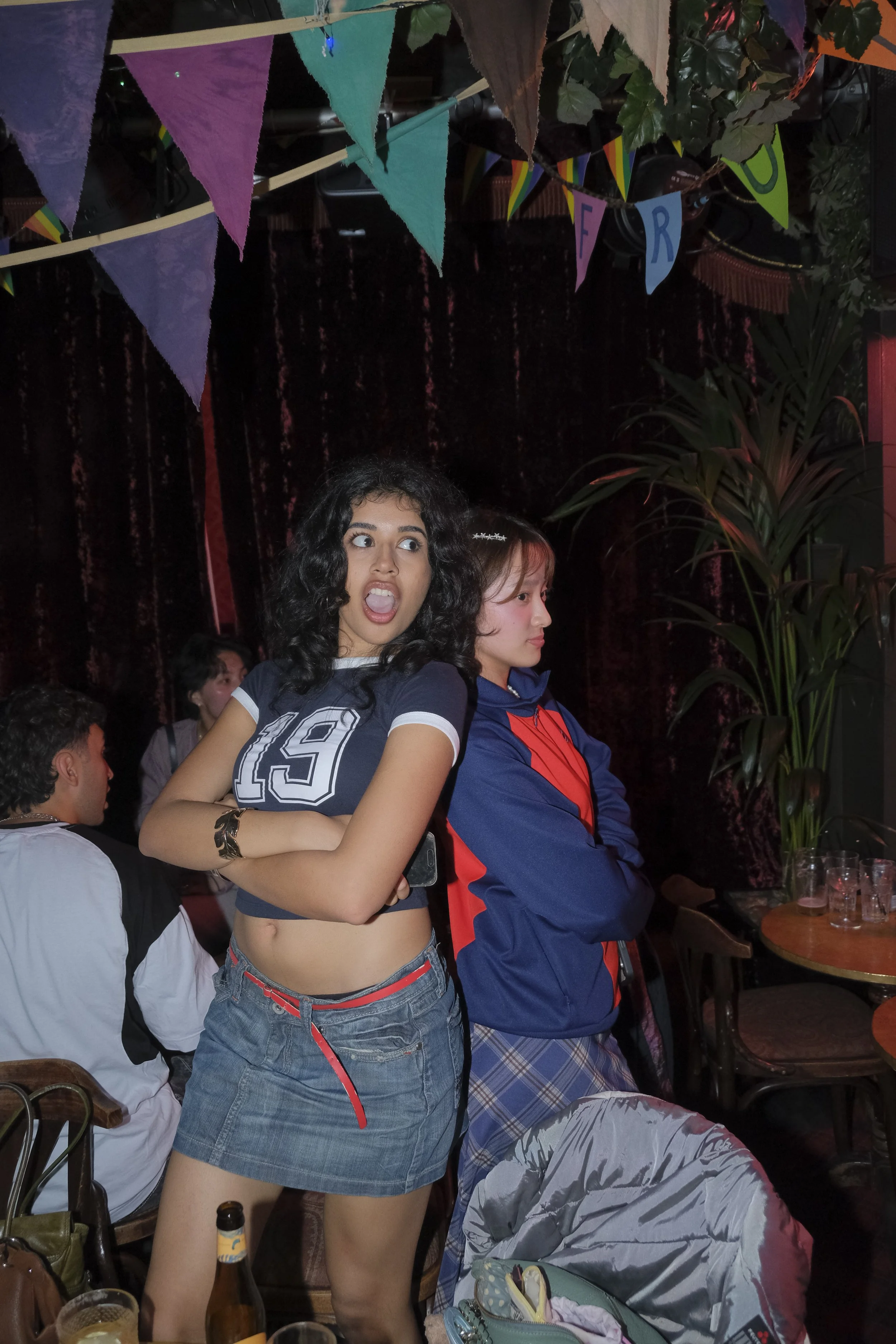 Two young women standing close together at a party or gathering, one with curly hair and expressive face, the other with straight hair and crossed arms, in a dimly lit room with colorful bunting and plants.