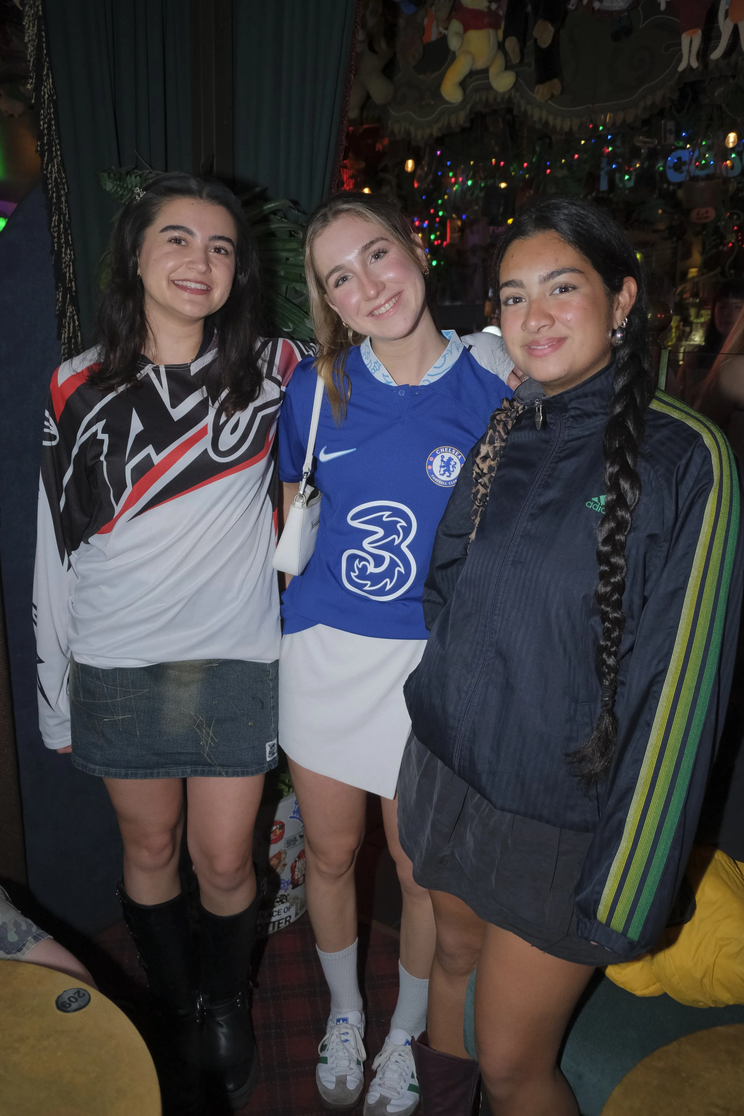 Three young women smiling and posing together indoors, with festive decorations and colorful lights in the background.