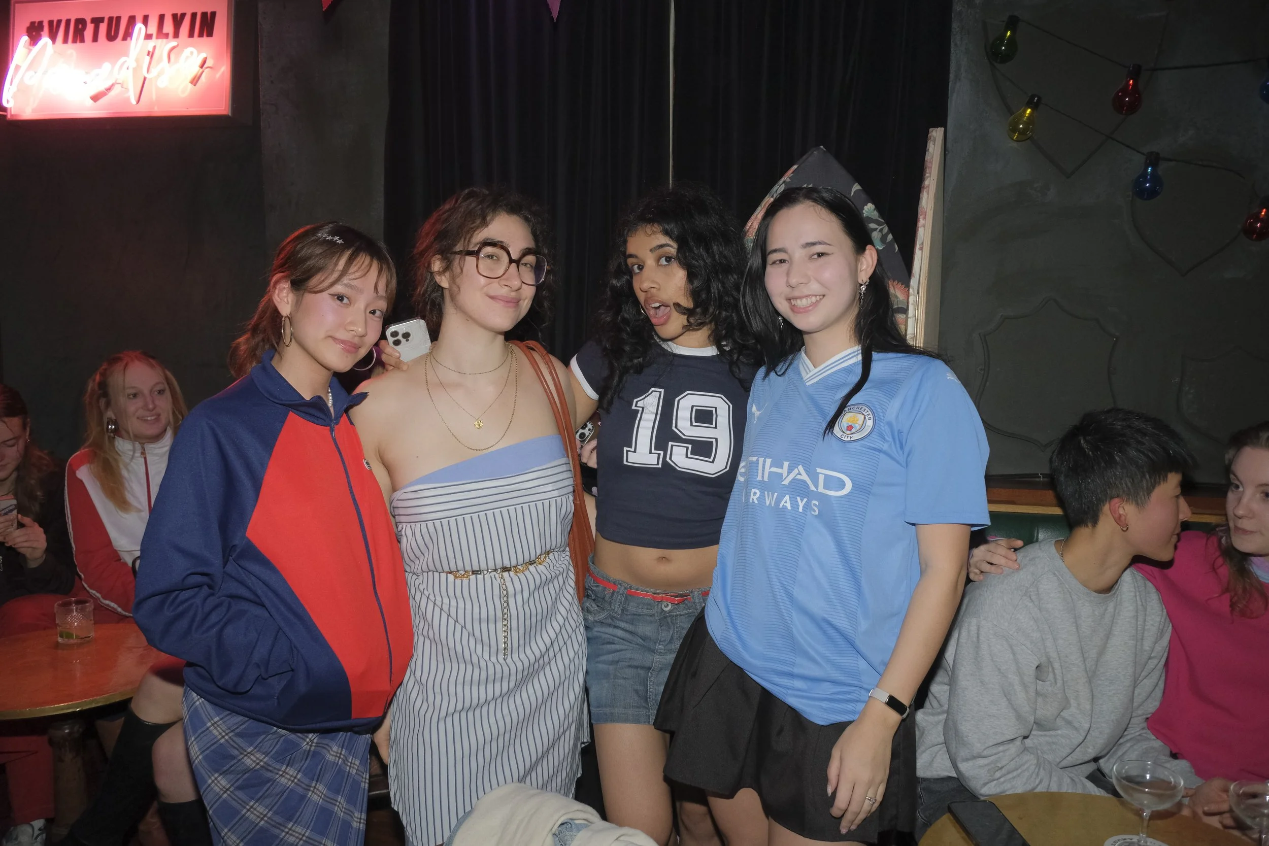 Group of young women posing together at a social gathering in a bar or club, some wearing casual and sports jerseys, with a neon sign in the background reading "VIRTUALLY LIN" and "Underground".