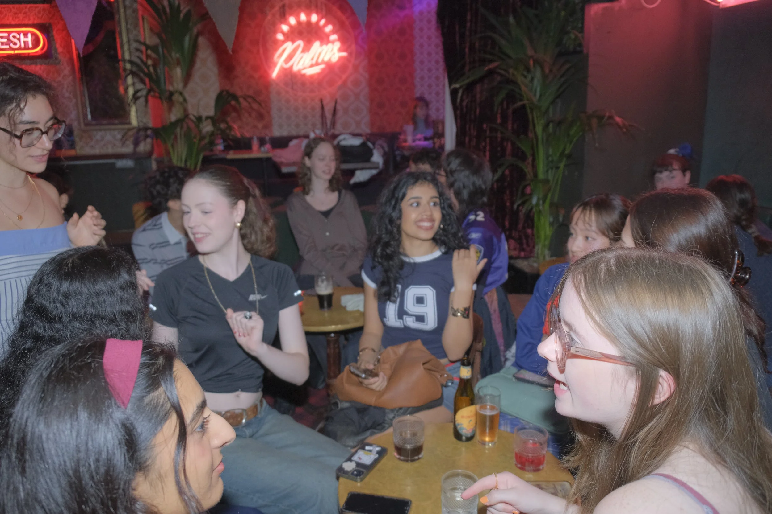 A group of young women socializing at a bar or nightclub with neon signs and plants in the background, some holding drinks and smiling.