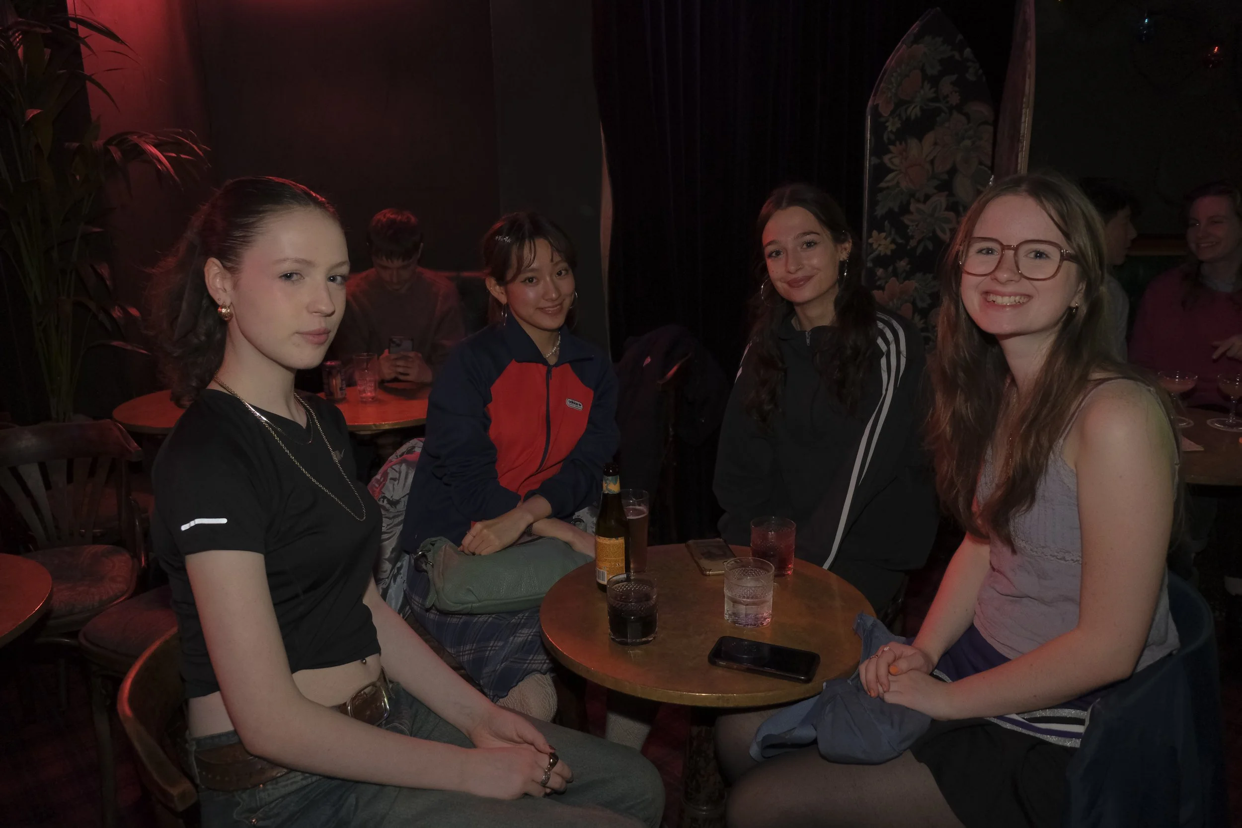 Four young women sitting at a round table in a dimly lit bar or restaurant, smiling, with drinks and a phone on the table.
