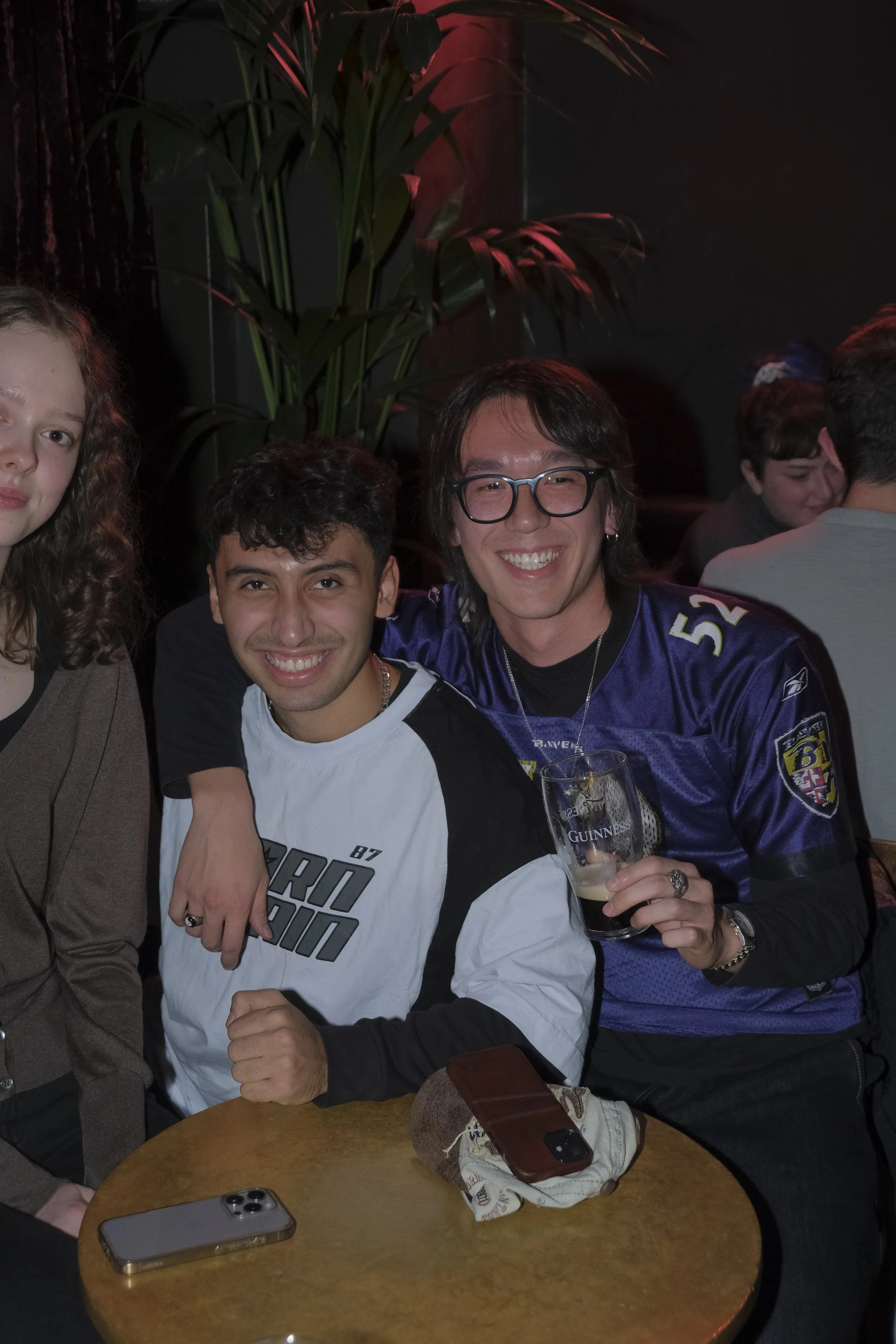 Three friends sitting at a table in a dimly lit bar or restaurant, smiling and enjoying drinks. One person is wearing a Baltimore Ravens football jersey, holding a pint of beer. The table has a smartphone, another phone, and a cloth with a cap. There