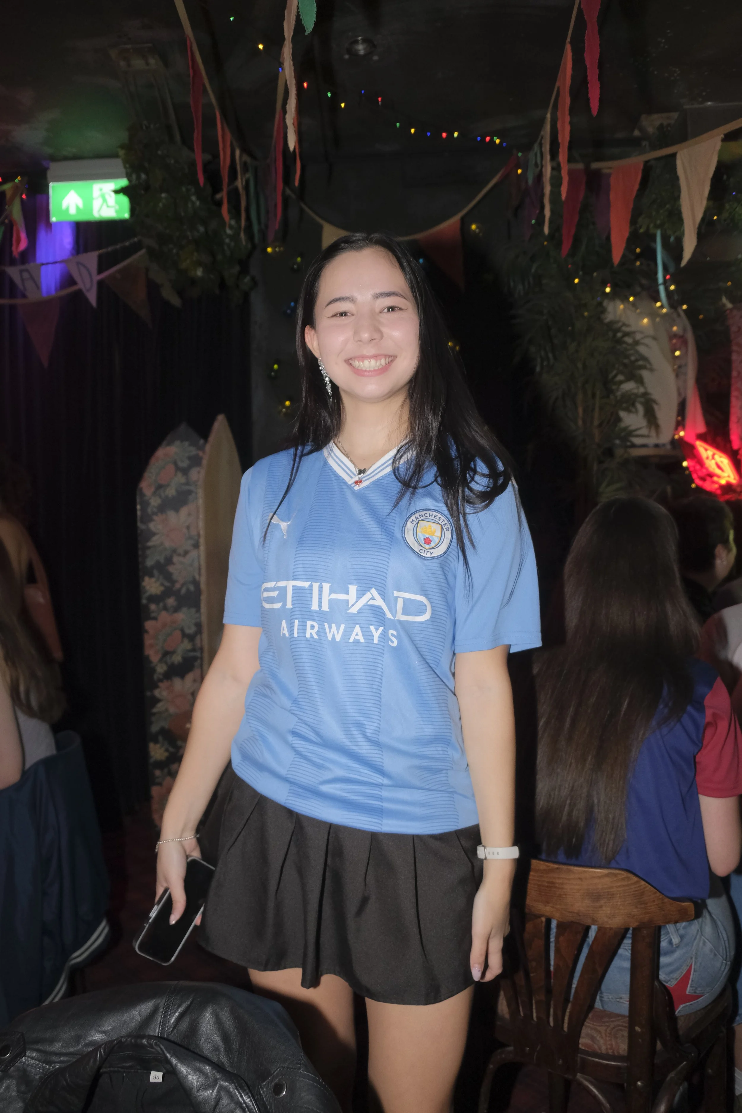 A young woman smiling and standing in a dimly lit room decorated with colorful banners and string lights. She is wearing a Manchester City football jersey and holding a smartphone in her left hand.