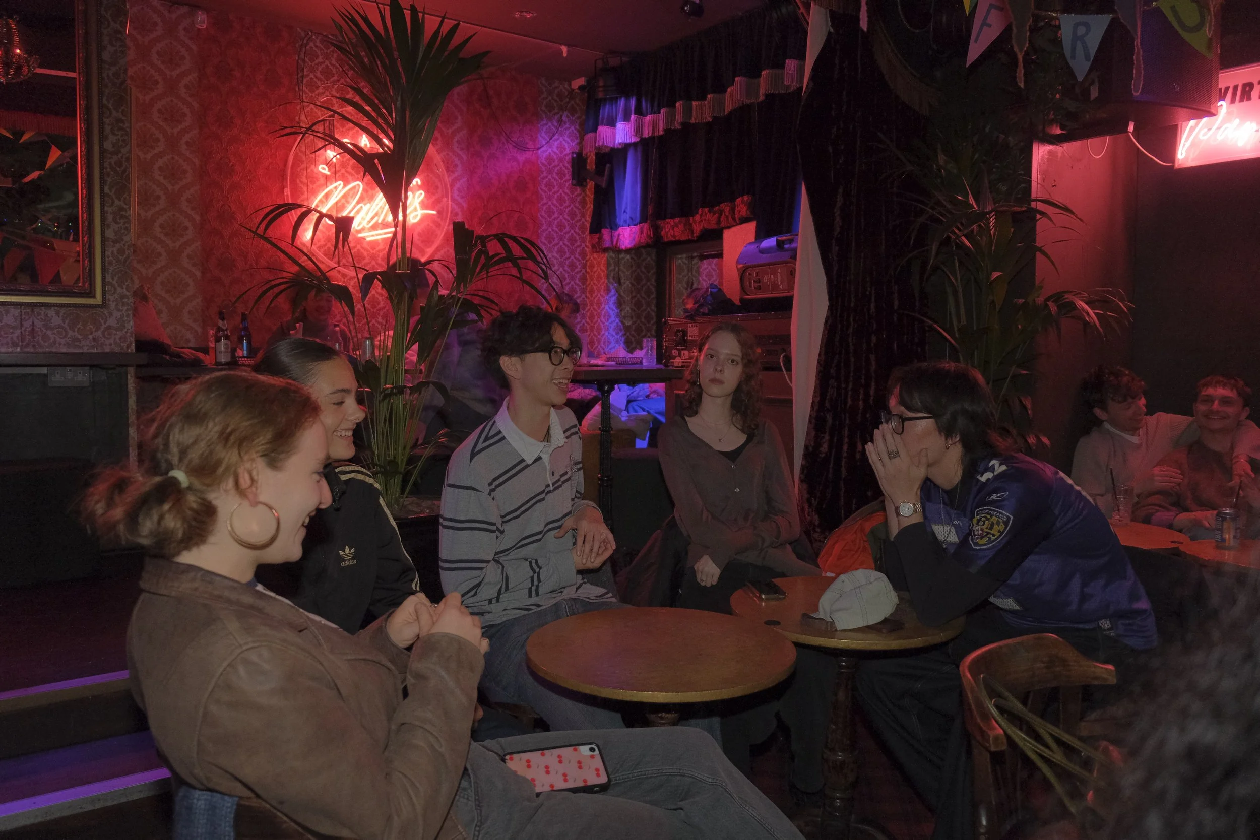 Group of people laughing and talking in a dimly lit bar or club with neon signs and plants.