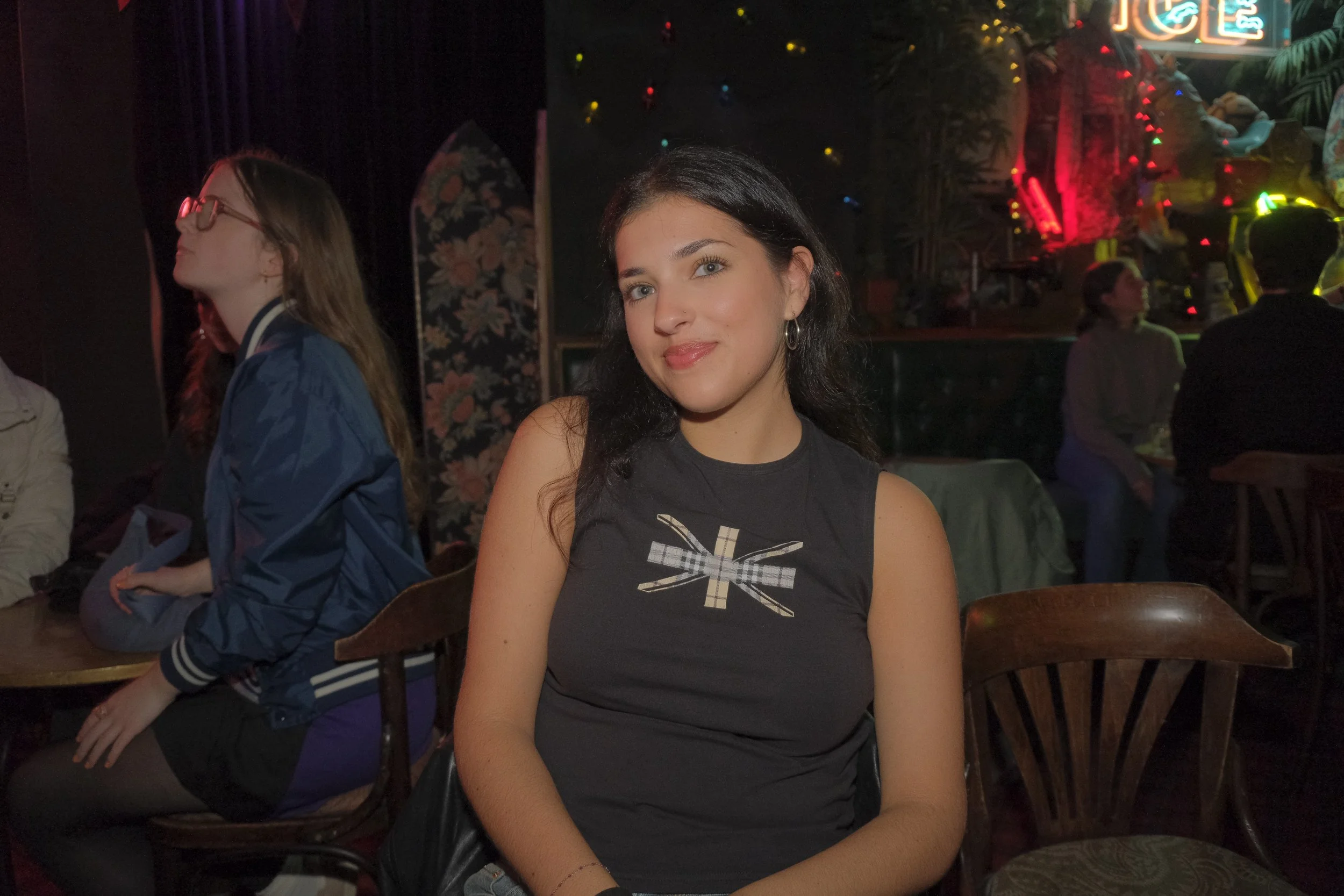Young woman with dark hair and hoop earrings sitting at a table in a dimly lit, festive setting with colorful decorations and other people in the background.