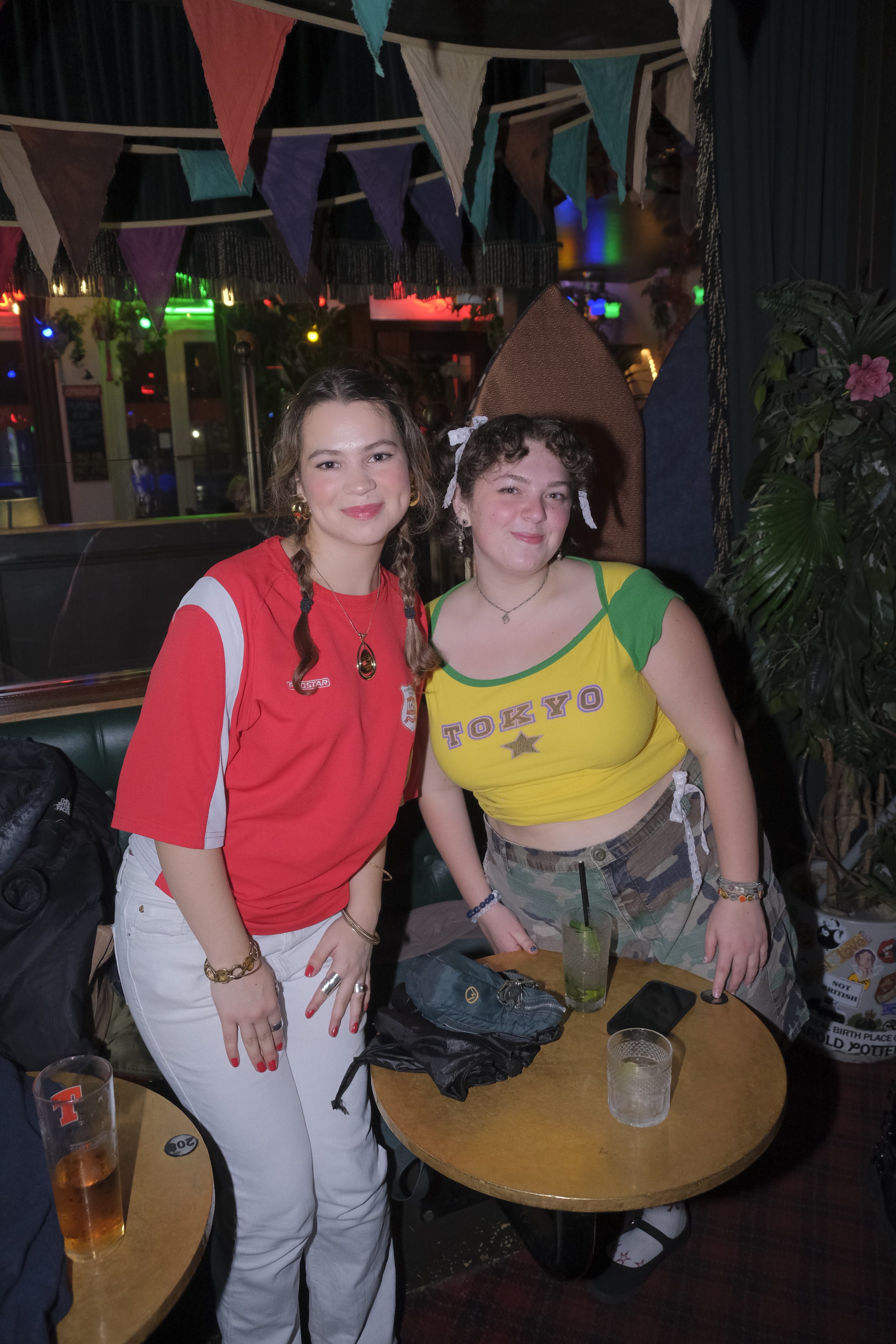 Two young women posing together at a bar or restaurant; one in a red sports shirt and white pants, the other in a yellow crop top and camouflage shorts, with colorful bunting hanging above.