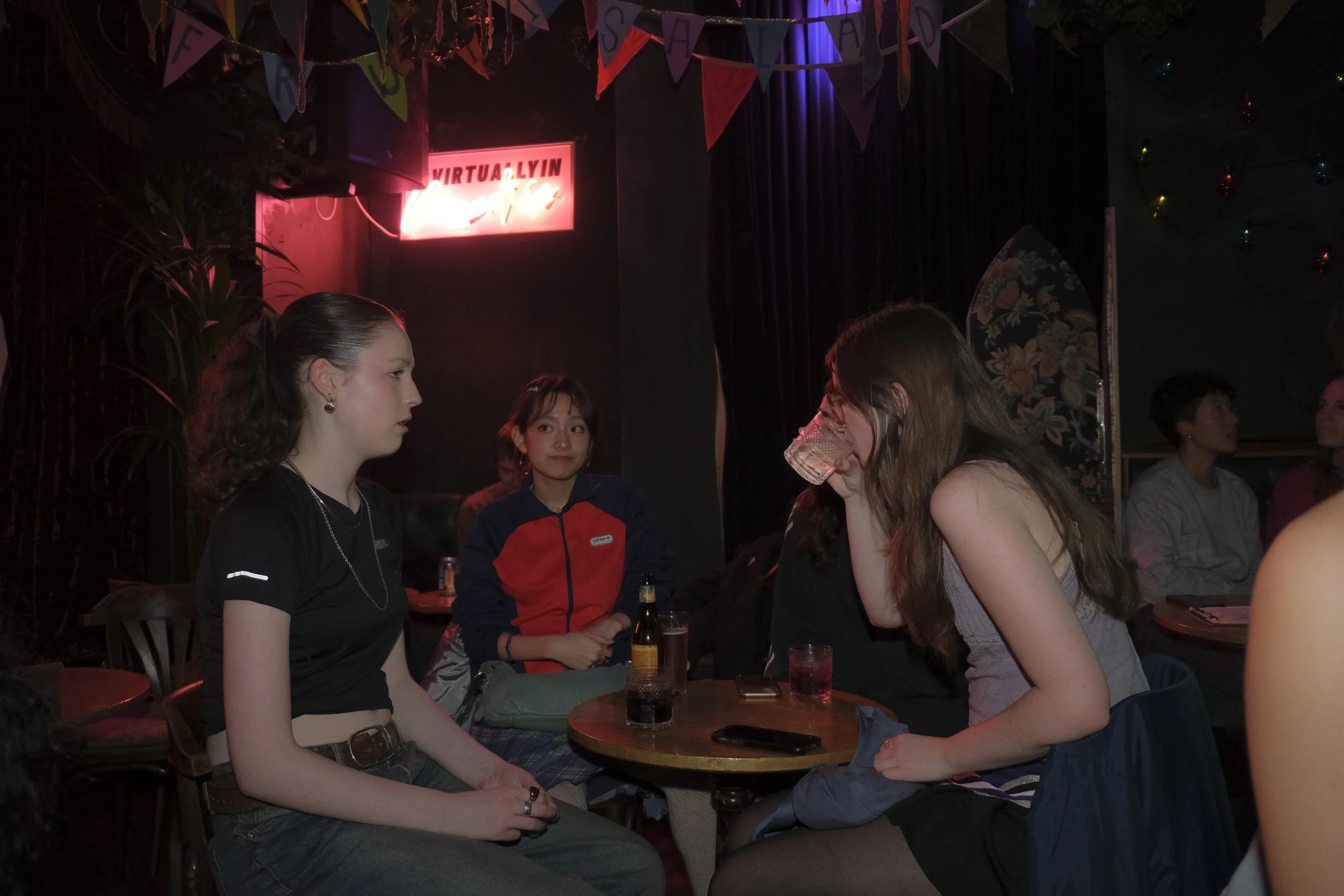 Four women are sitting around a small round table in a dimly lit bar or club with a neon sign and colorful flags hanging overhead. One woman on the right is drinking from a glass, while the others are engaged in conversation.