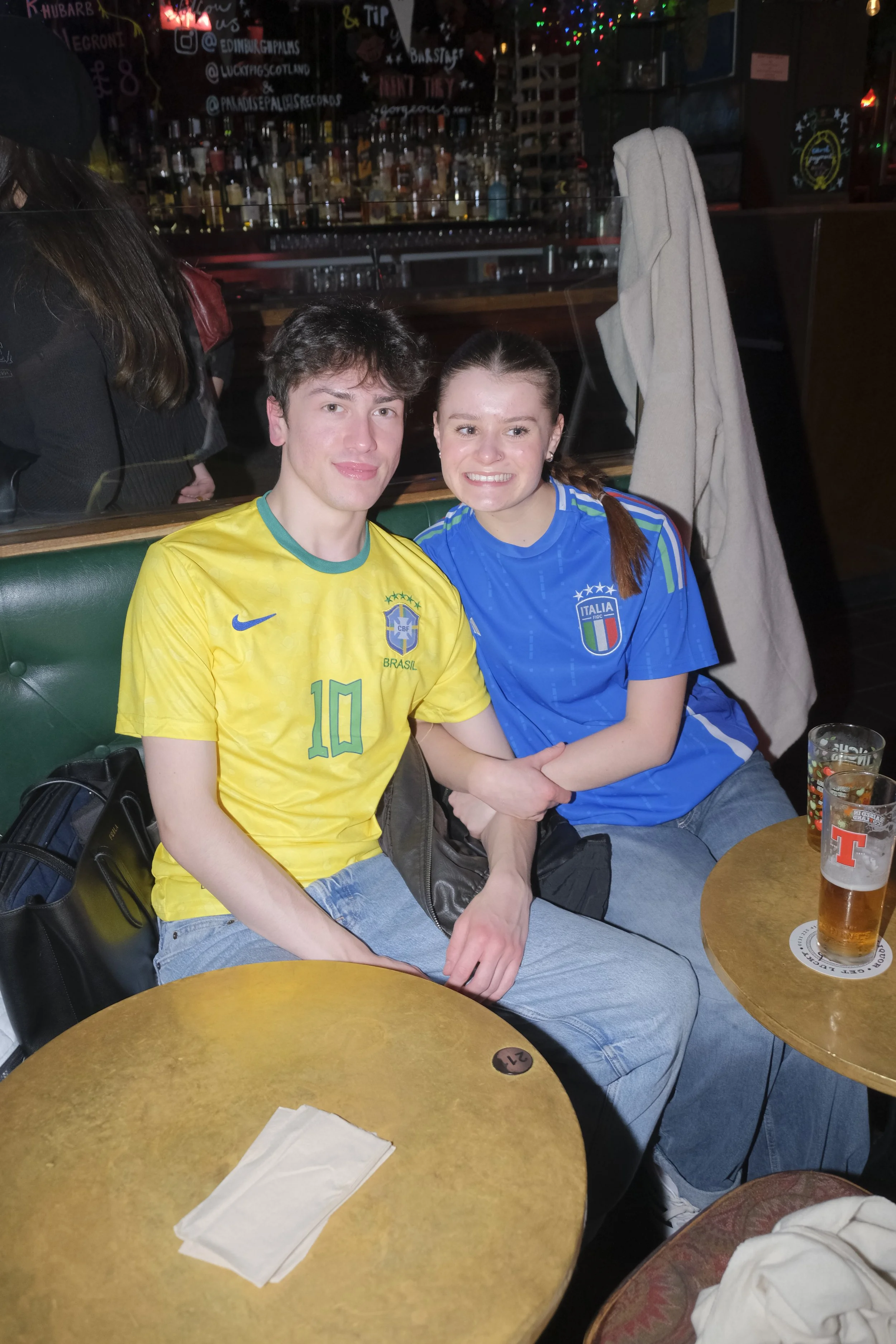 Two young adults, a man and a woman, sitting at a bar, wearing Brazil and Italy soccer jerseys respectively, smiling and holding hands. There are drinks on the table and a bar with bottles in the background.