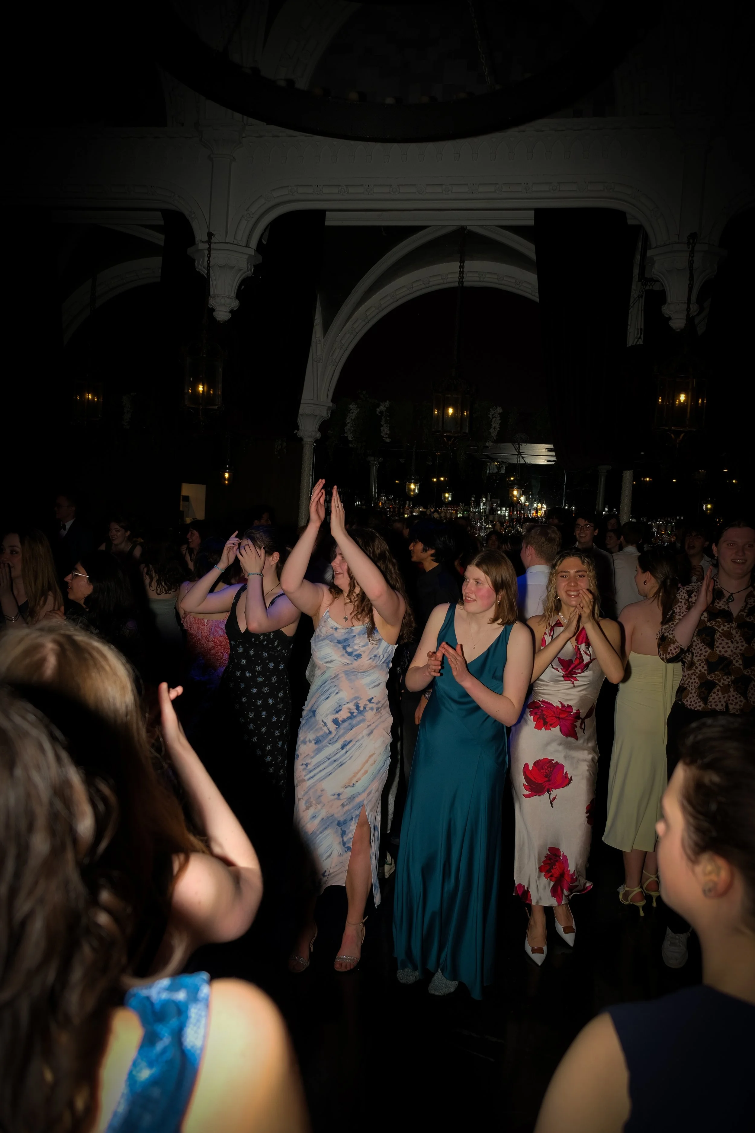 People dancing and celebrating at an indoor event, some with hands raised in the air, wearing colorful dresses and formal attire.