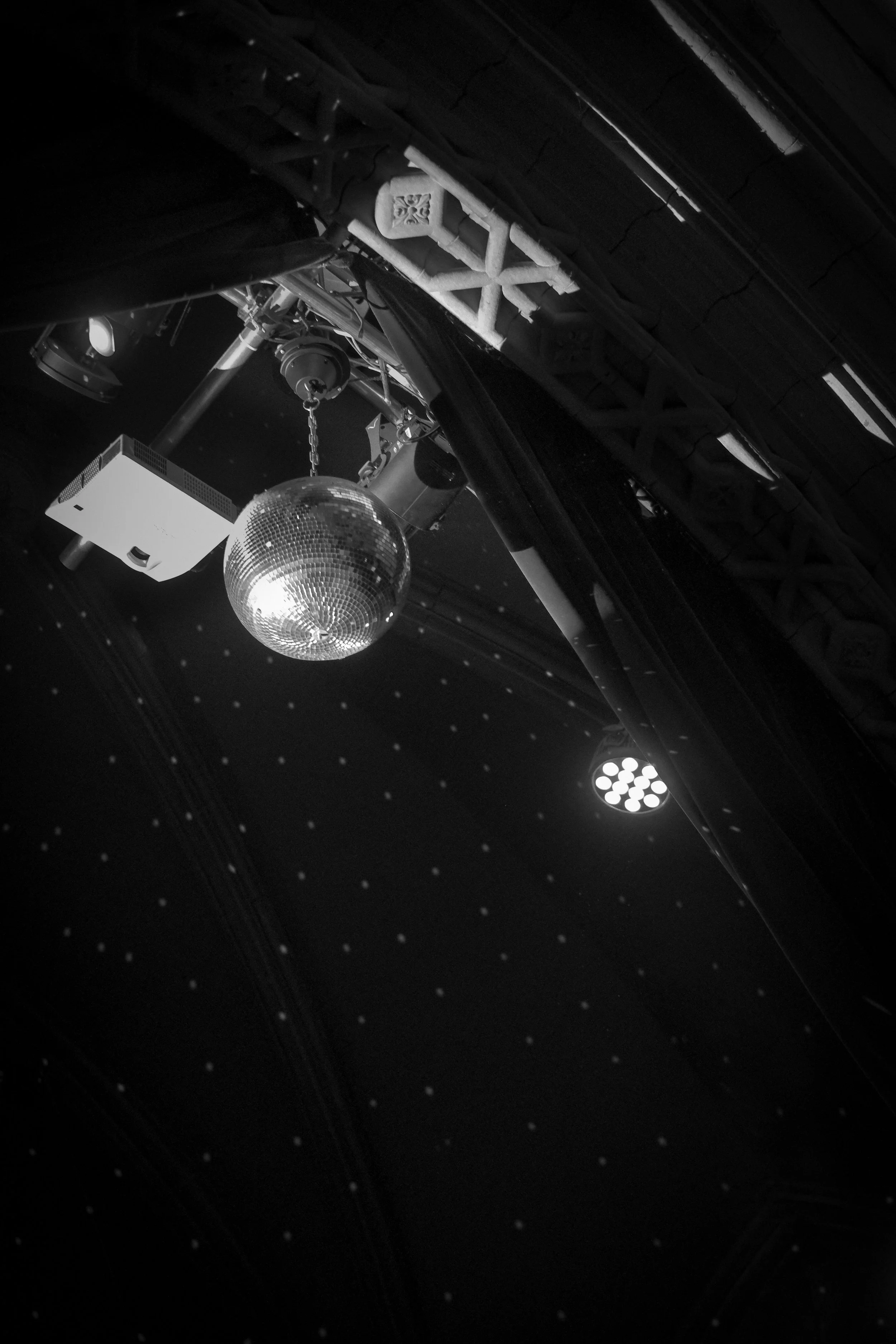 A black-and-white photograph of a ceiling with a disco ball, stage lights, and curtains, suggesting a nightclub or performance venue.
