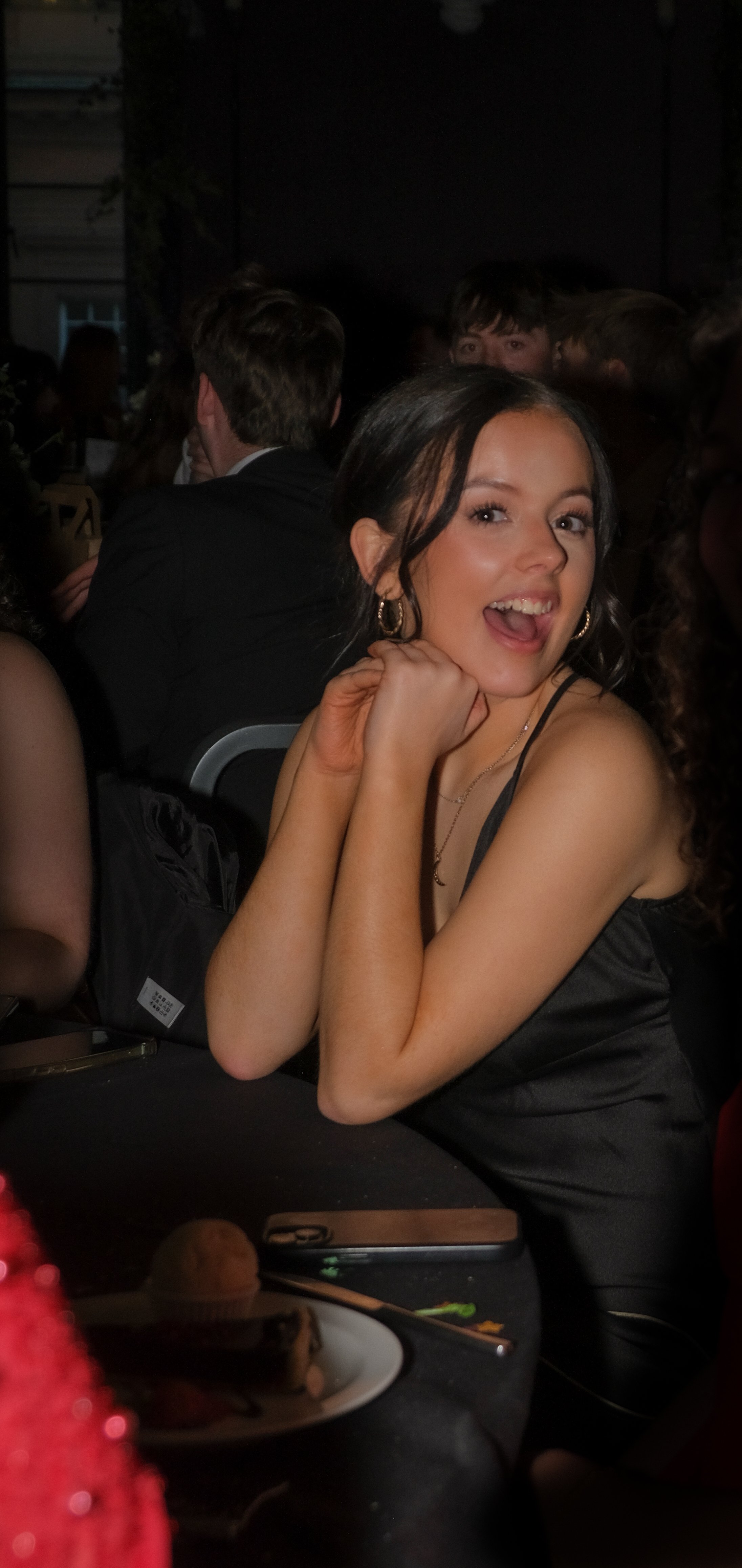 A young woman with shoulder-length dark hair, wearing gold hoop earrings and a black spaghetti-strap dress, sitting at a table in a dimly lit indoor setting, smiling and resting her chin on her hand.