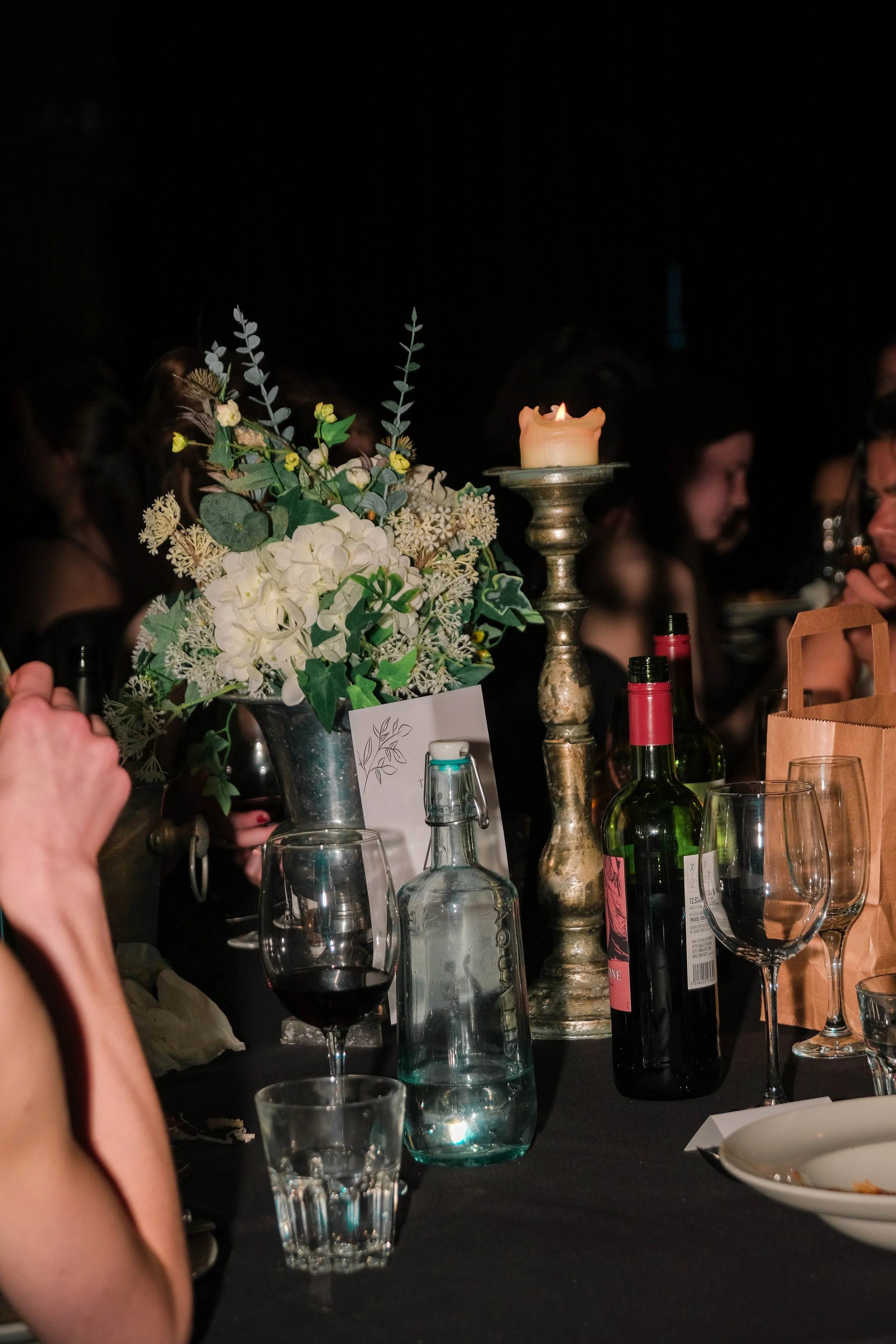 A table decorated with a floral arrangement, a lit candle on a candlestick, wine bottles, glasses, and plates at a dinner event, with people in the background.