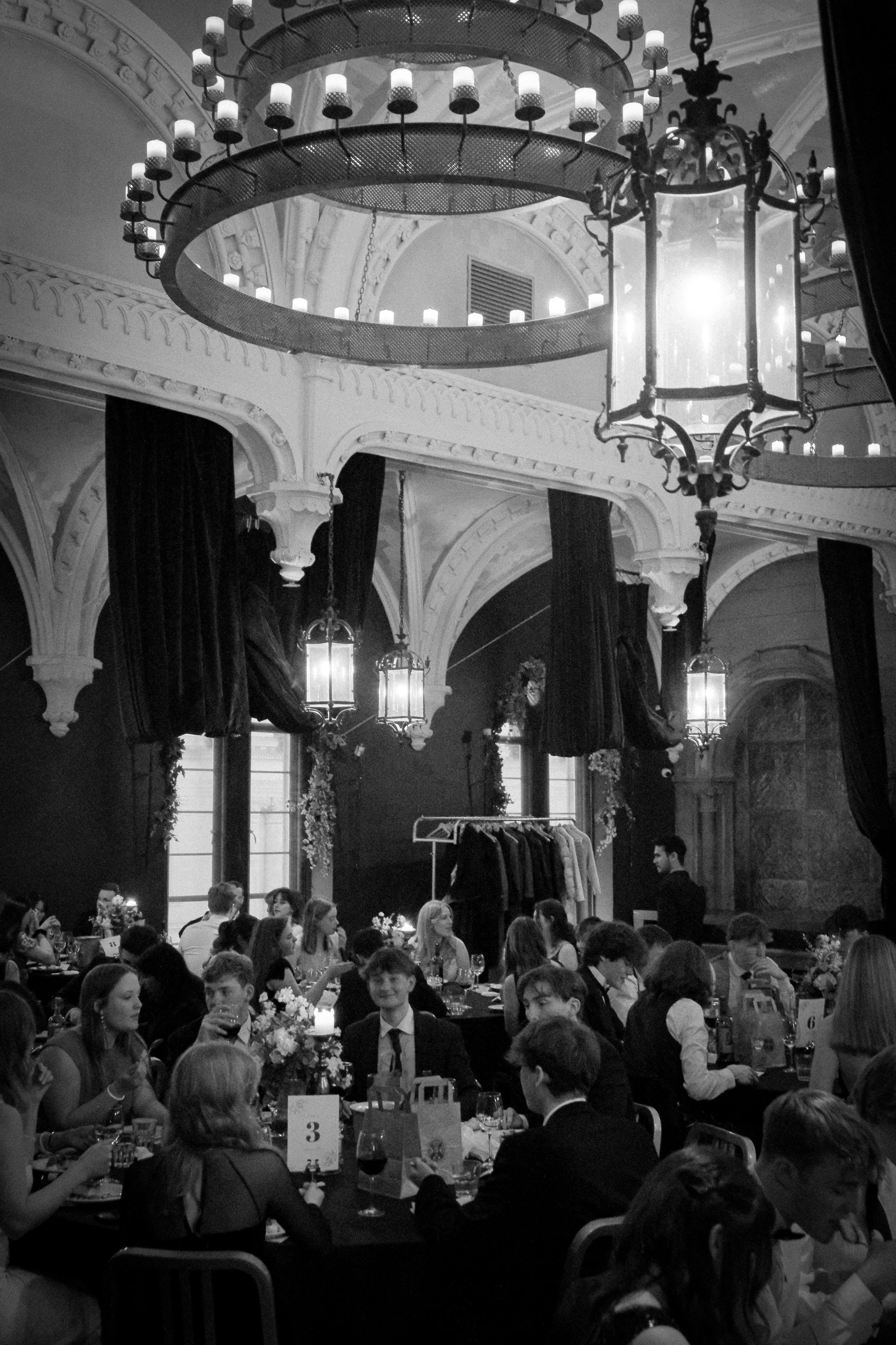 An indoor party scene in a decorated hall with multiple guests sitting at round tables, some with drinks, and a coat rack with clothing. The hall has large windows, dark curtains, ornate architectural details, and large chandeliers hanging from the c