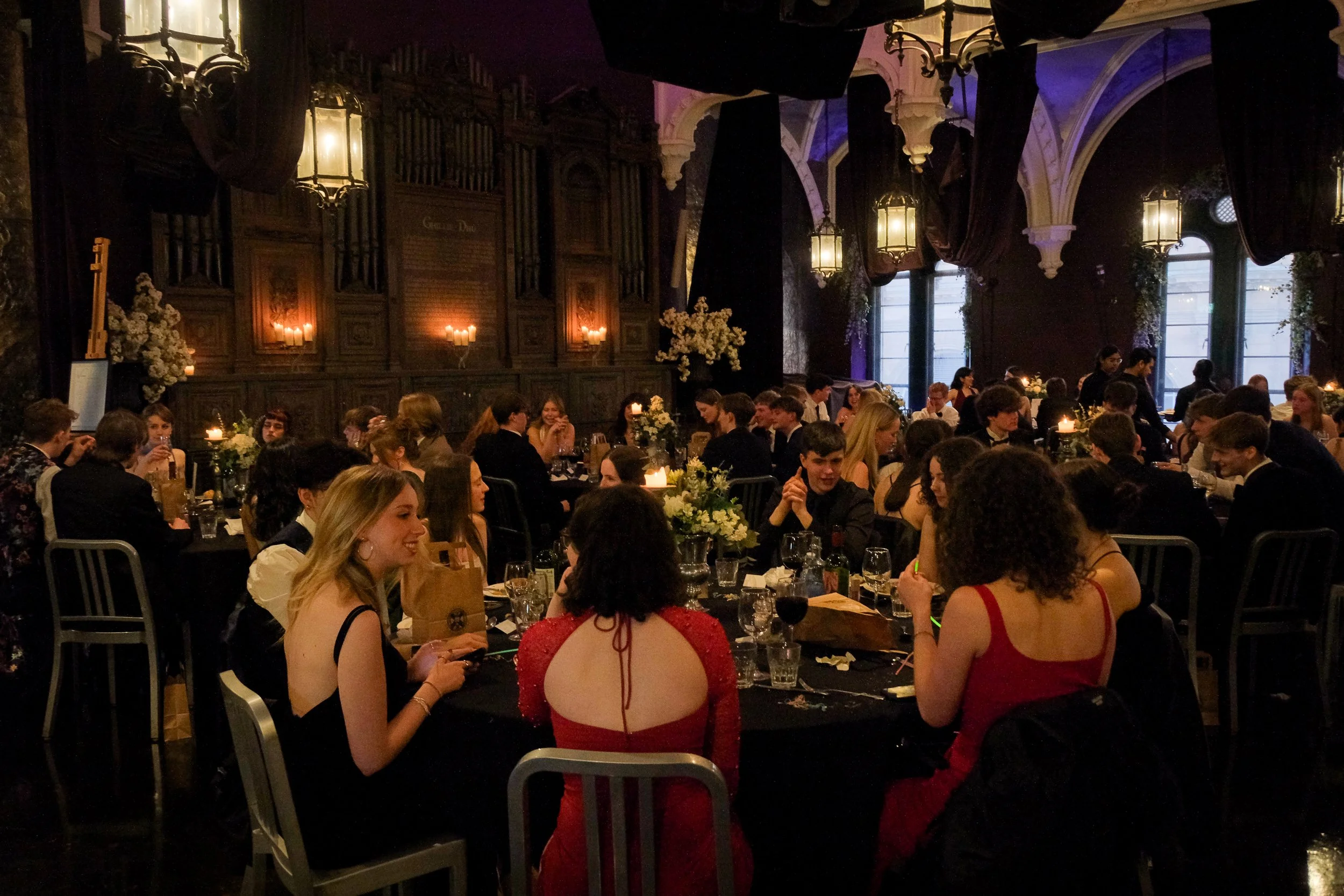 People sitting around decorated tables in a dimly lit banquet hall with dark wood paneling, floral centerpieces, and hanging lantern-style chandeliers.