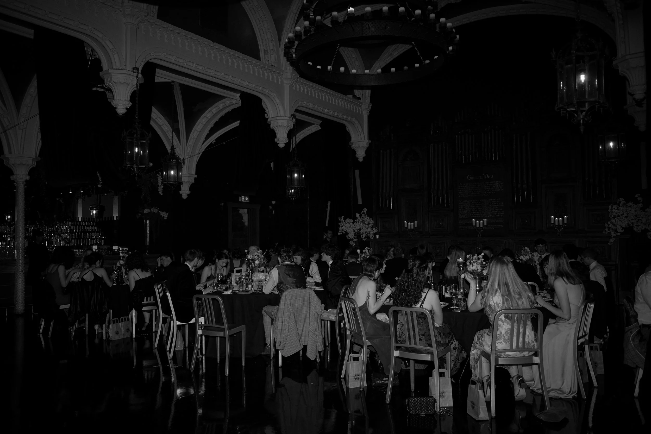 A formal dinner event with numerous people seated around a long table in an ornate hall with high, arching ceilings and chandeliers, dim lighting, and floral decorations.