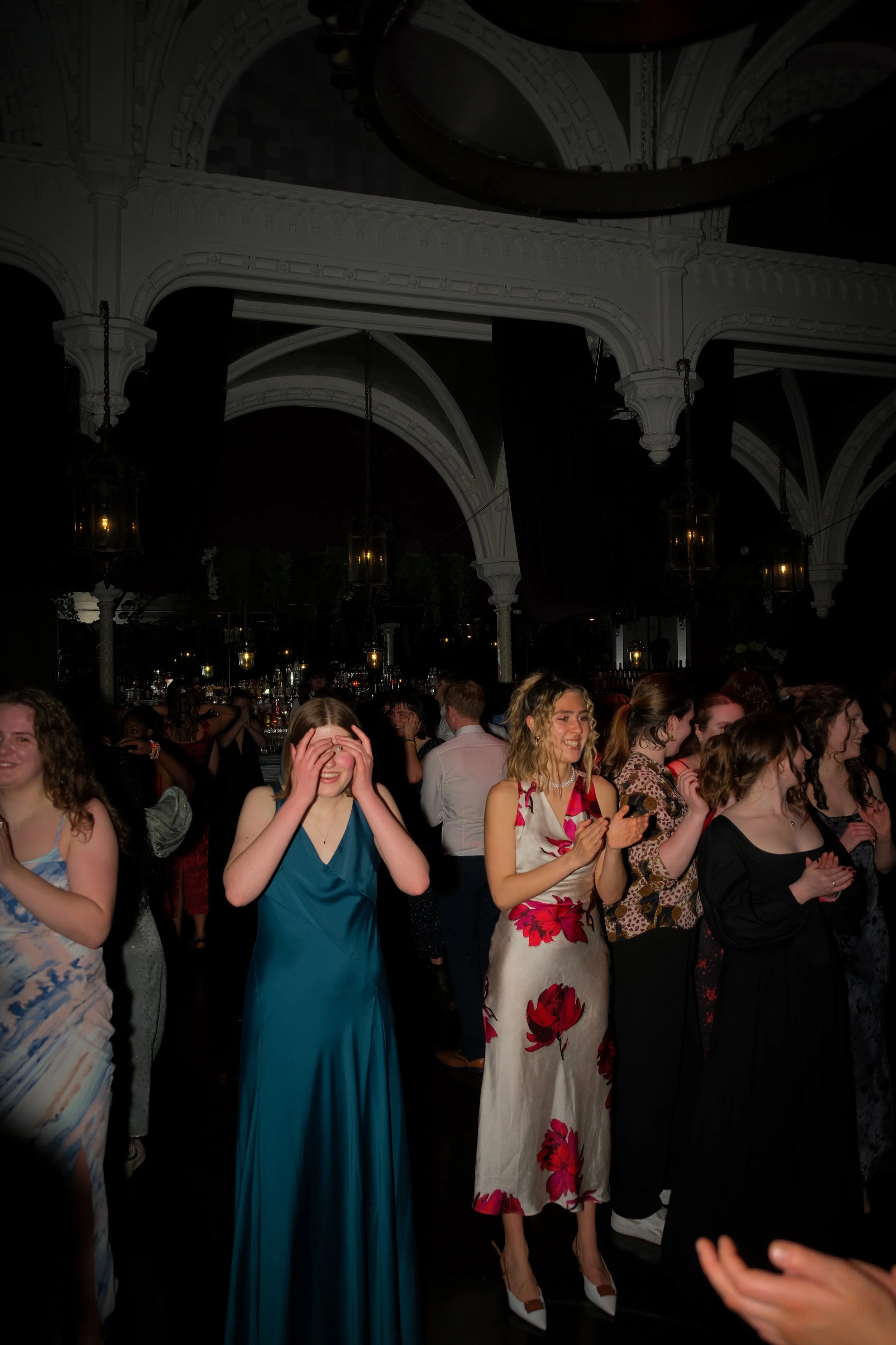 Group of young women at a dance or party, some smiling and clapping, in an elegant venue with decorative arches and hanging lanterns.
