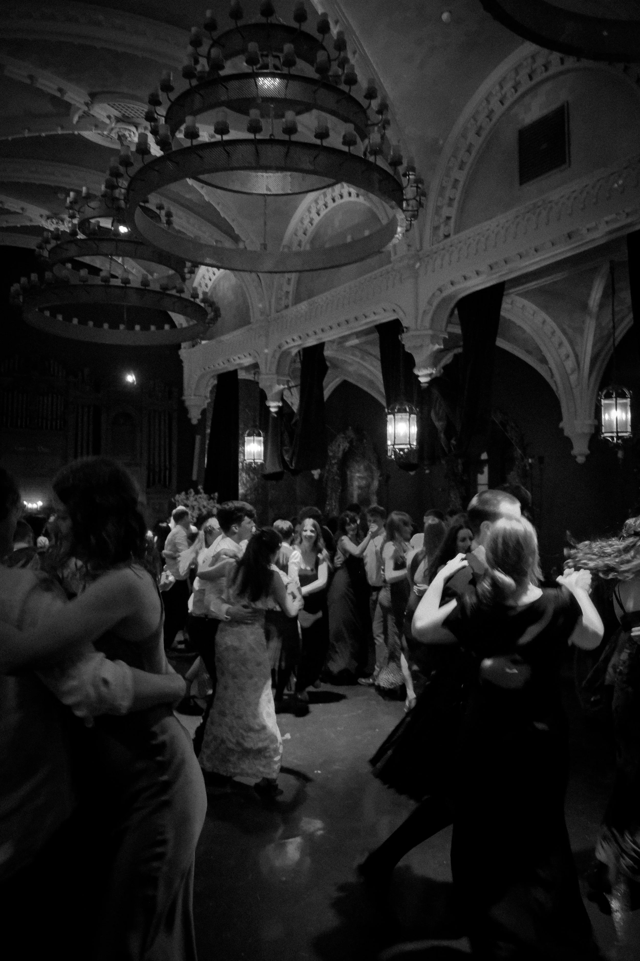 People dancing in a dimly lit ballroom with ornate architecture and large chandeliers.