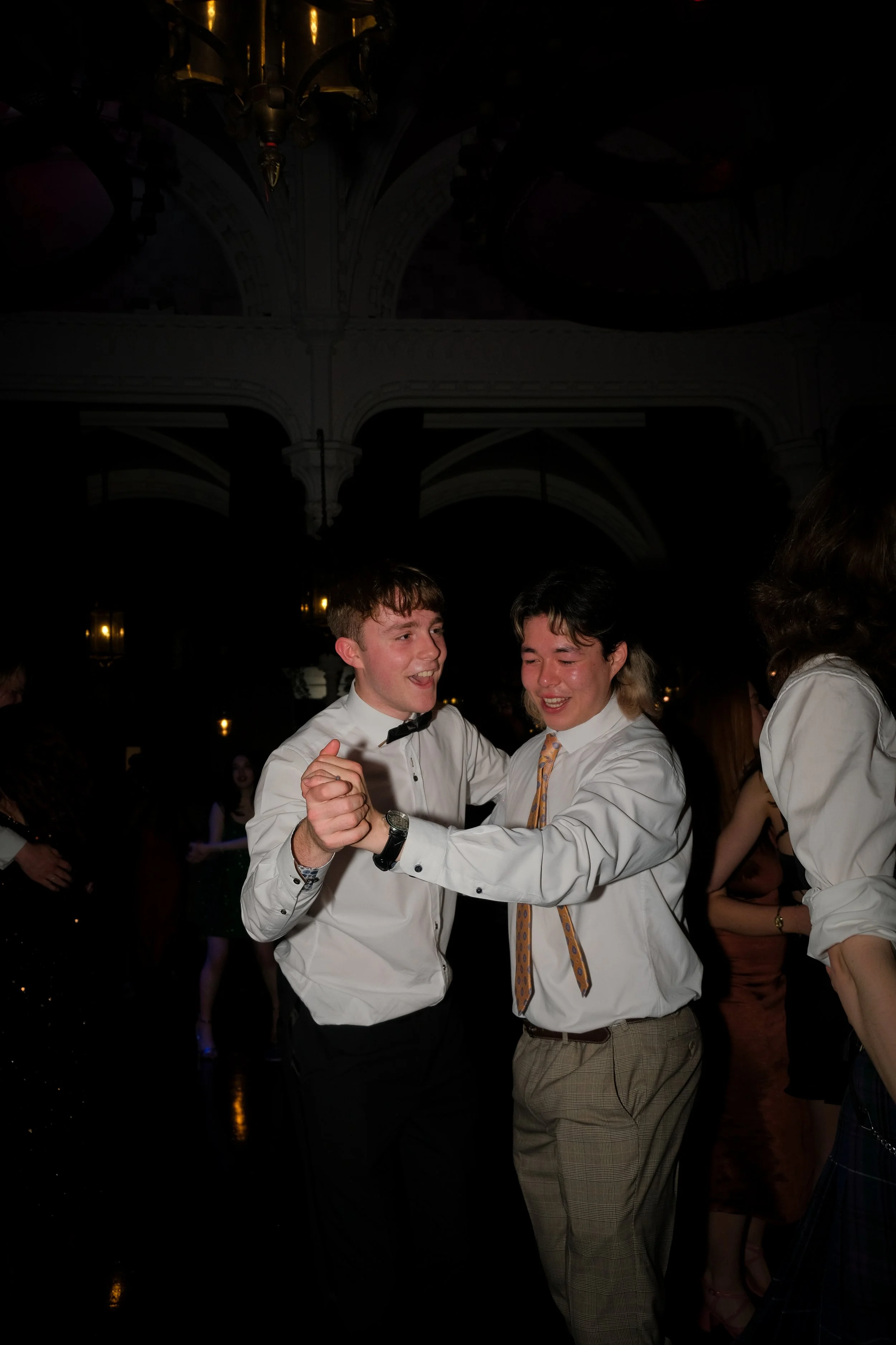 Two young men dancing happily at a formal event or party. One is wearing a white shirt with a bow tie, the other is wearing a white shirt with a patterned tie. They are smiling and holding hands as they dance.