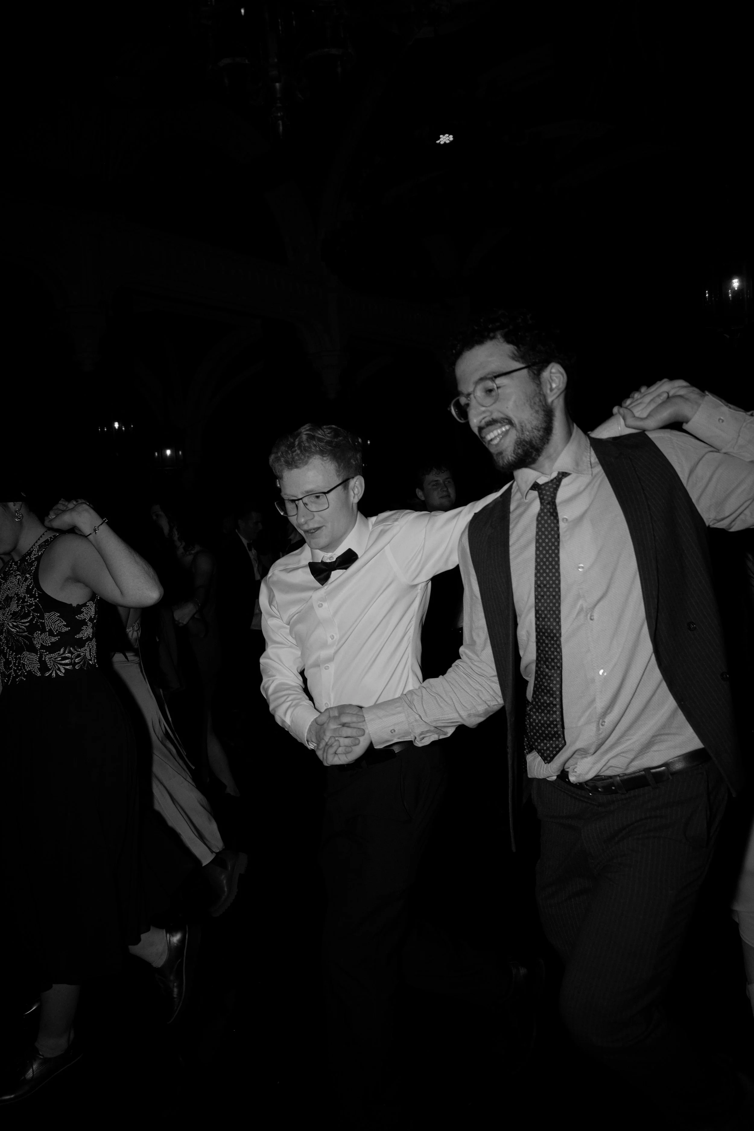 Two men, one wearing a bow tie and the other a tie, holding hands while dancing at a formal event, with others dancing in the background.