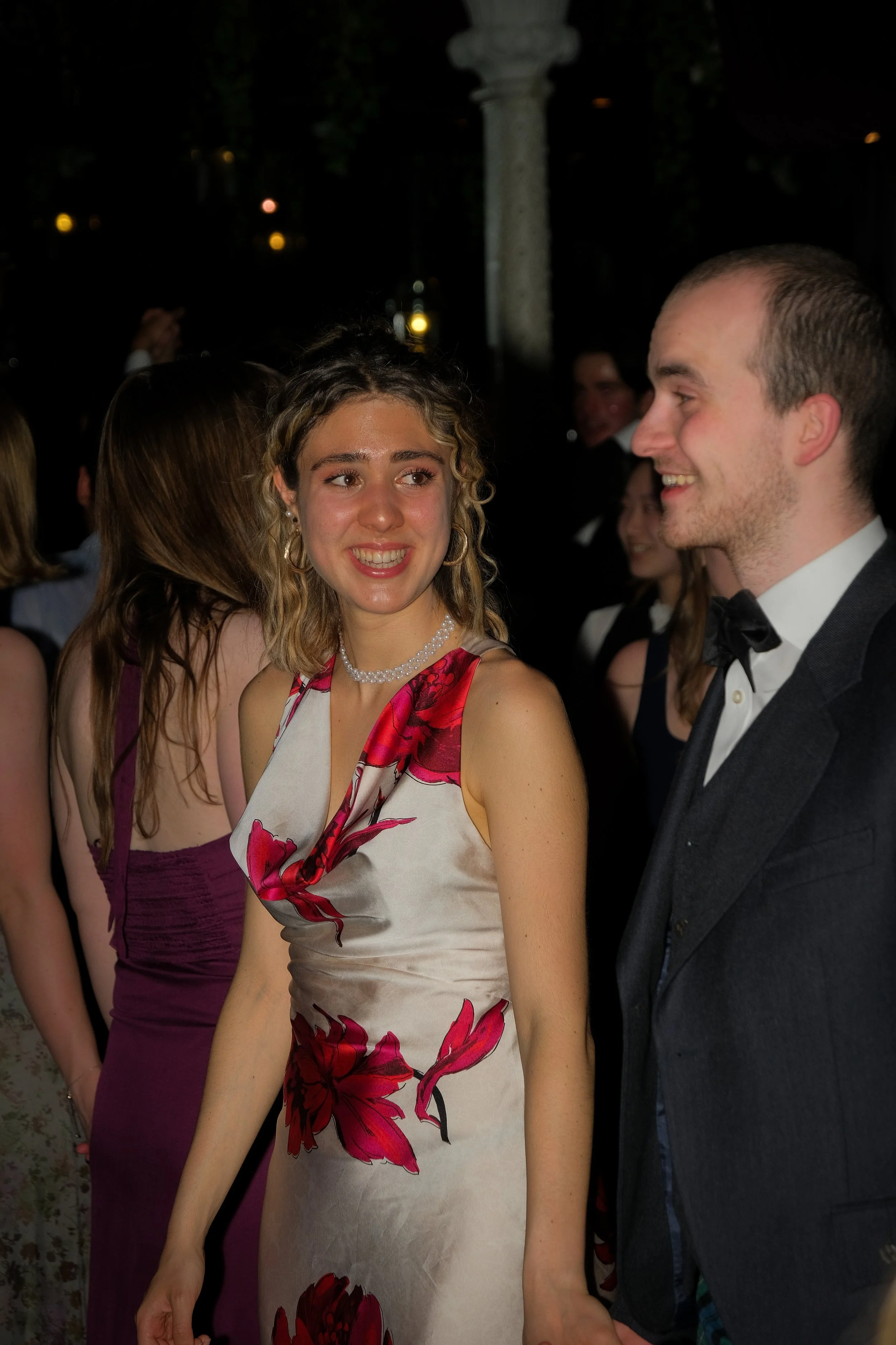 A young woman with curly blonde hair in a floral dress and pearl necklace smiling at a young man in a tuxedo at a formal event.