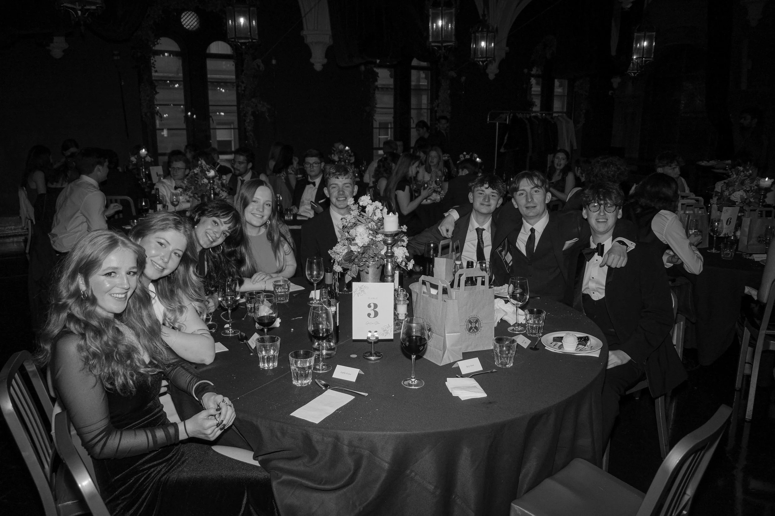 Group of young people sitting at a round table during a formal event or dinner, smiling at the camera, with glasses, plates, and a floral centerpiece on the table.