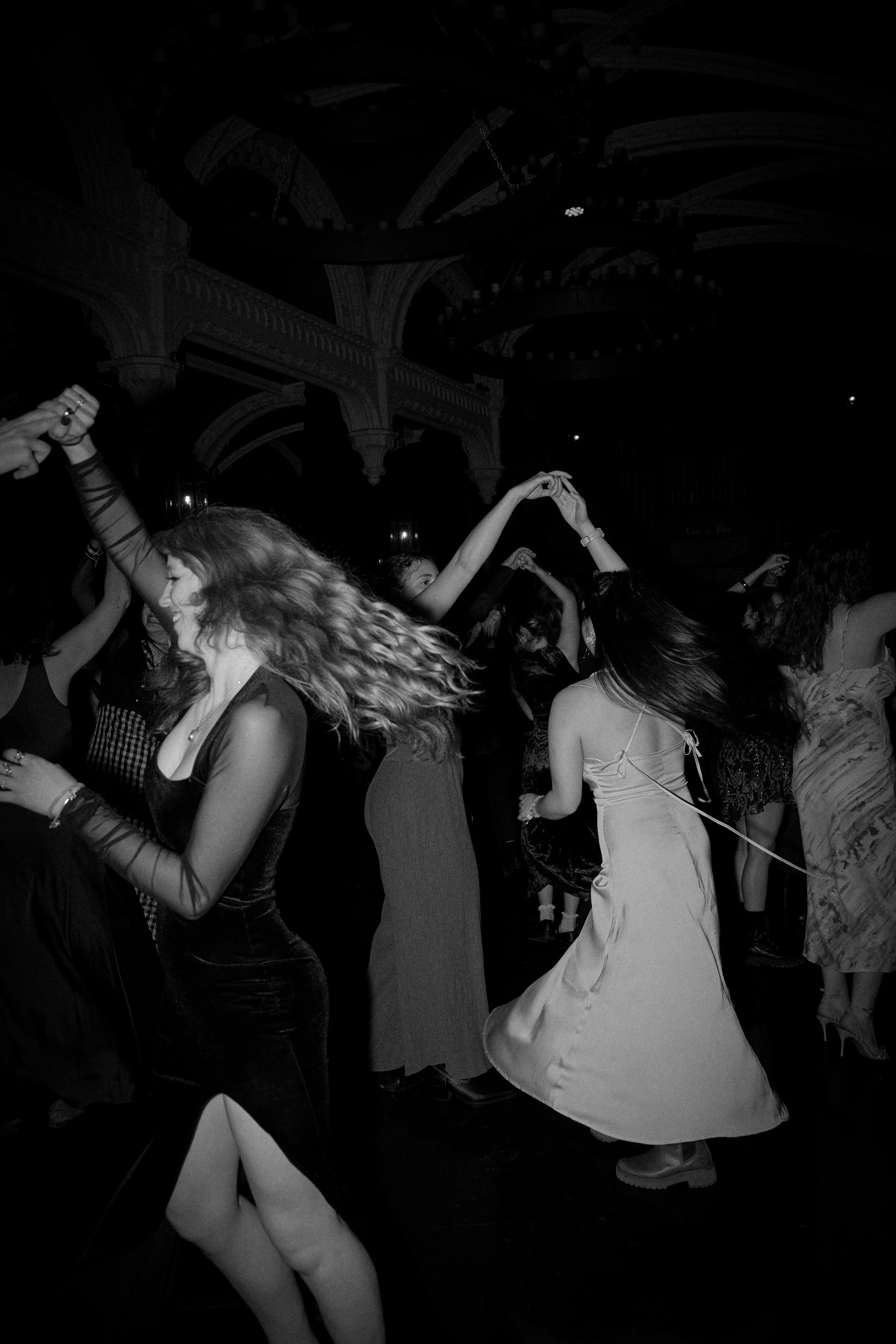 People dancing at a party or nightclub, with their hands in the air and hair flowing, under ornate ceiling architecture.