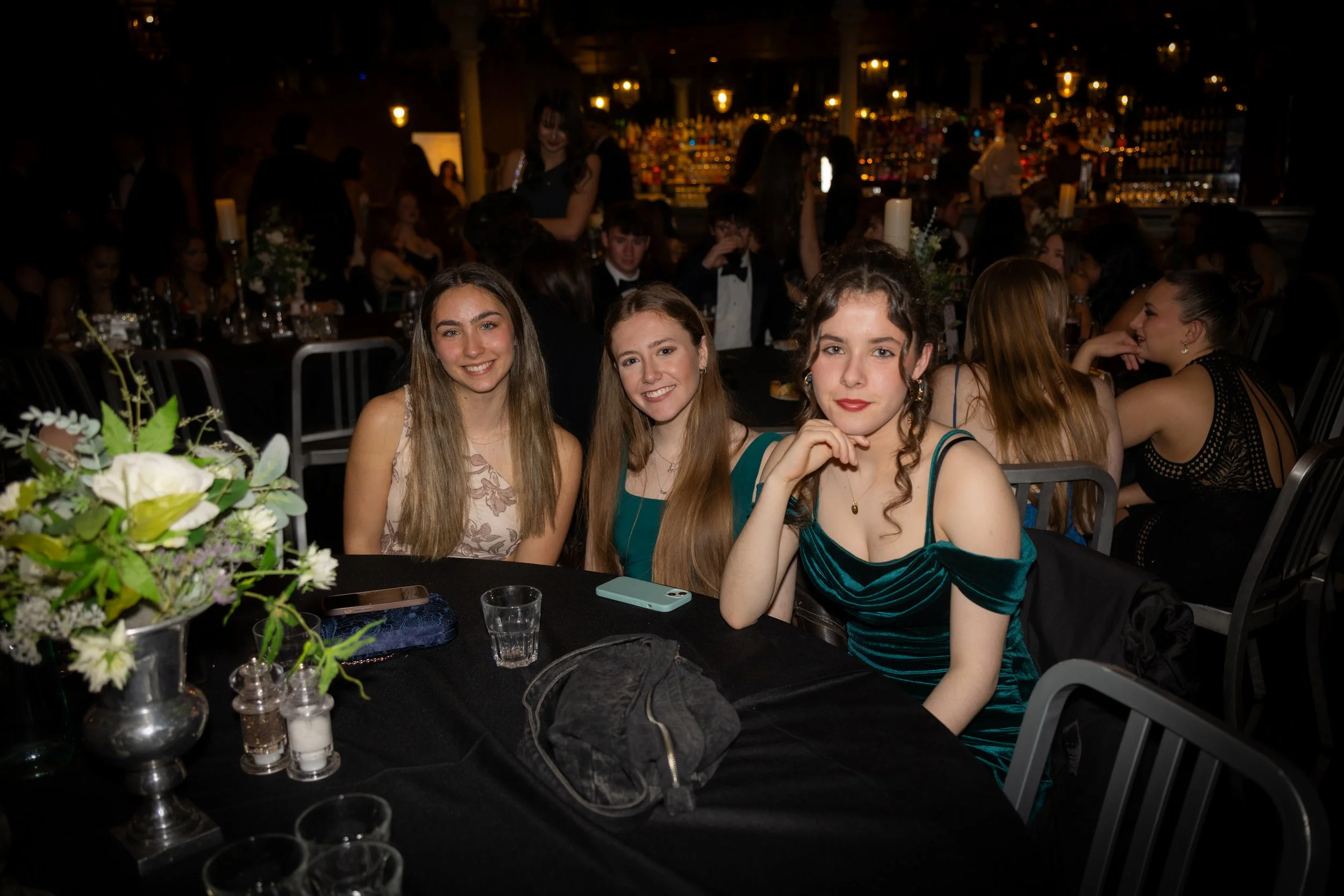 Three young women sitting at a decorated table during a formal event, with a social gathering and bar in the background.