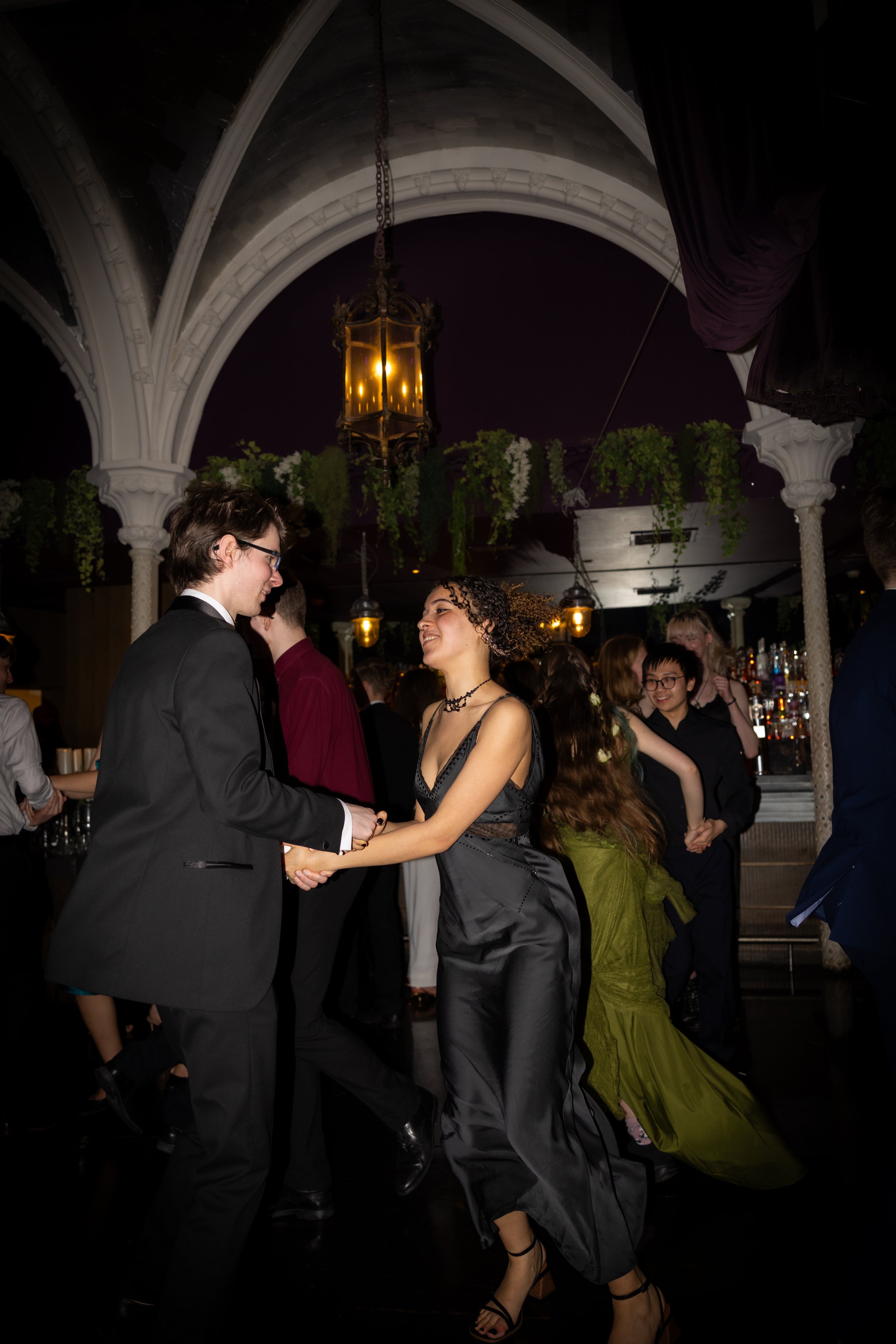 People dancing at a formal event, with men in tuxedos and women in elegant dresses, under ornate arches and hanging lanterns.