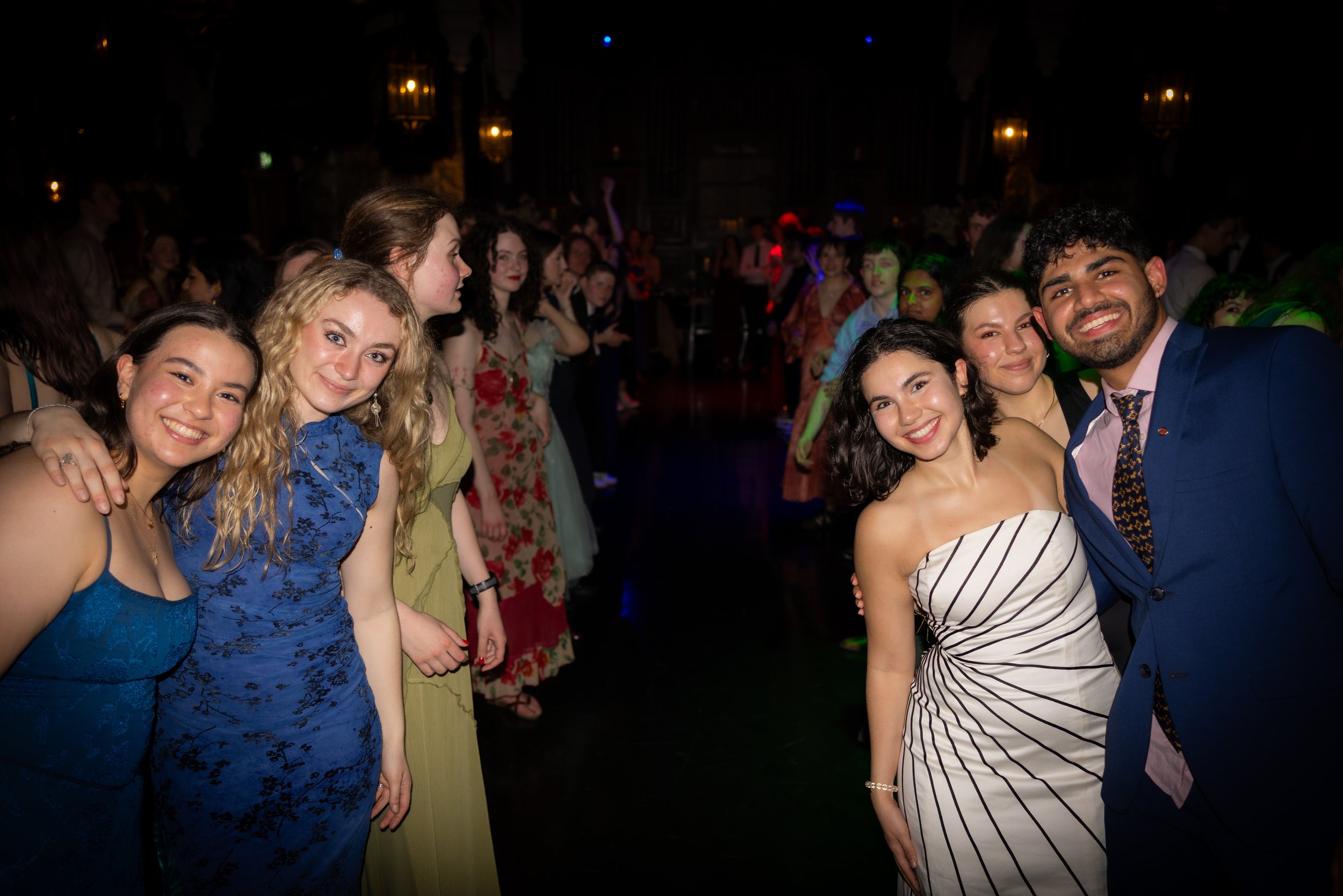 Group of young people at a party or celebration, smiling and posing for a photo in a dimly lit venue with colorful lighting.