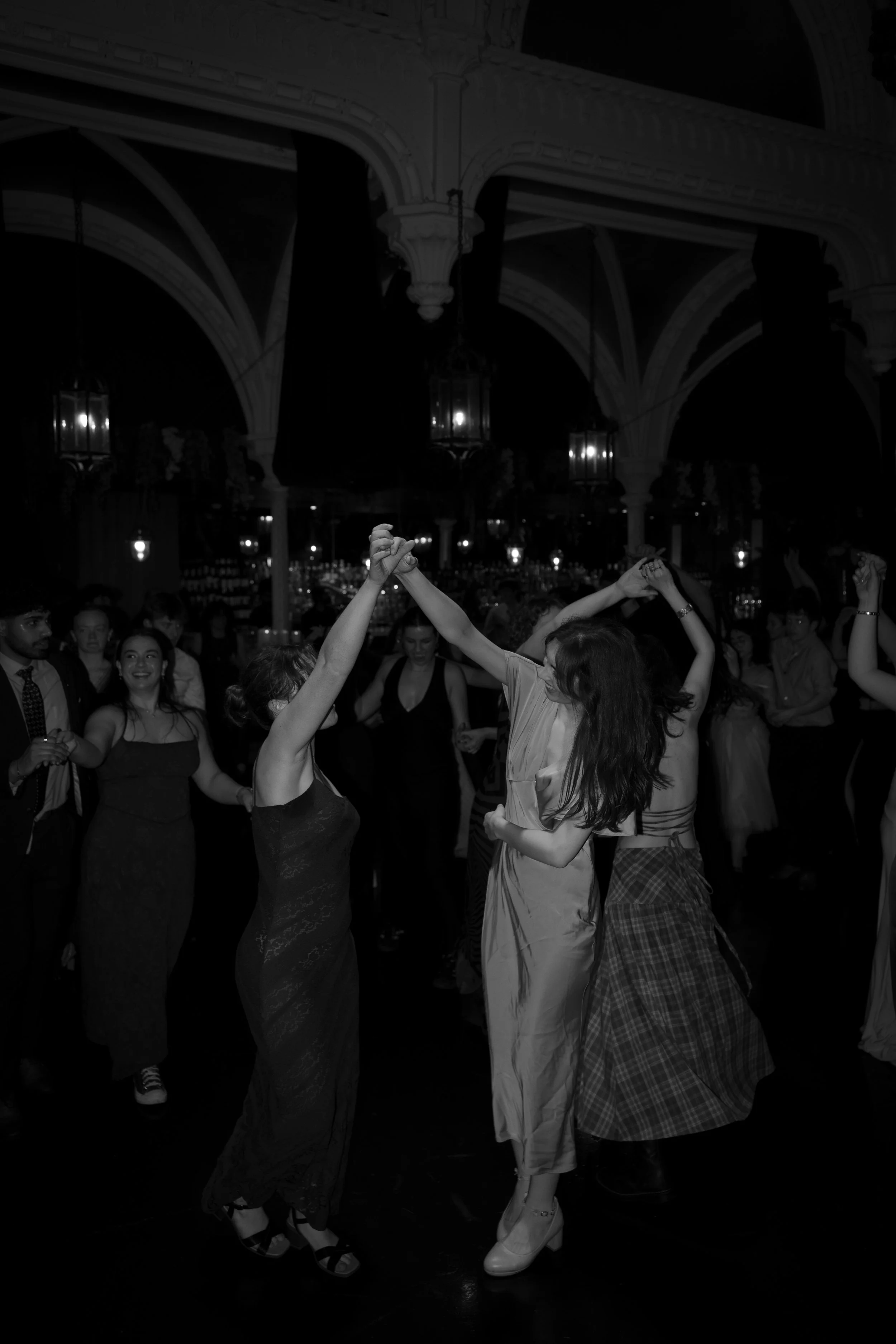 People dancing together in a dimly lit venue with high ceilings and chandeliers, smiling and holding hands.