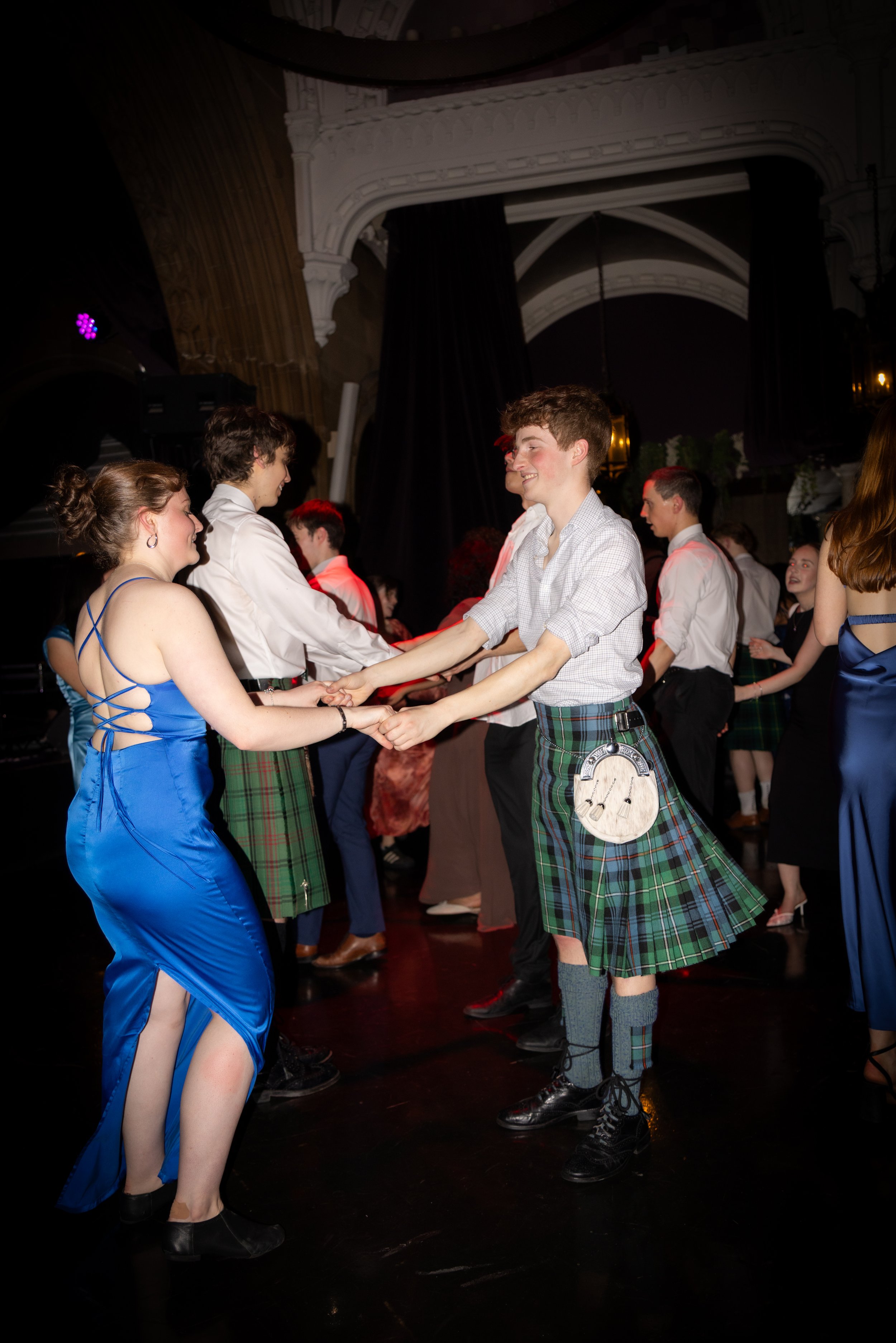 Young people dancing in formal attire at a dance event, with some wearing traditional Scottish kilts.