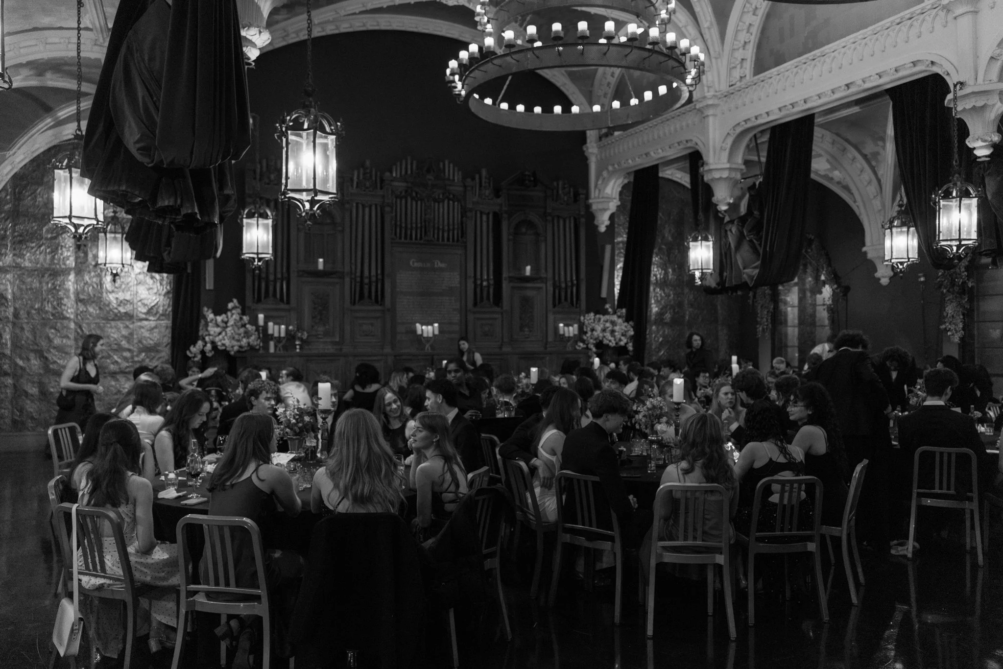 A large banquet hall filled with formally dressed guests seated at tables decorated with flowers and candles, with a stage at the front and ornate chandeliers hanging from the ceiling.