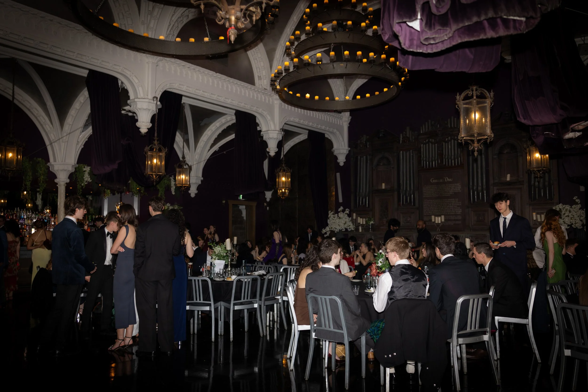 People dressed formally at a large, elegant banquet or wedding reception in a grand hall with ornate white arches, chandeliers, and hanging lanterns.