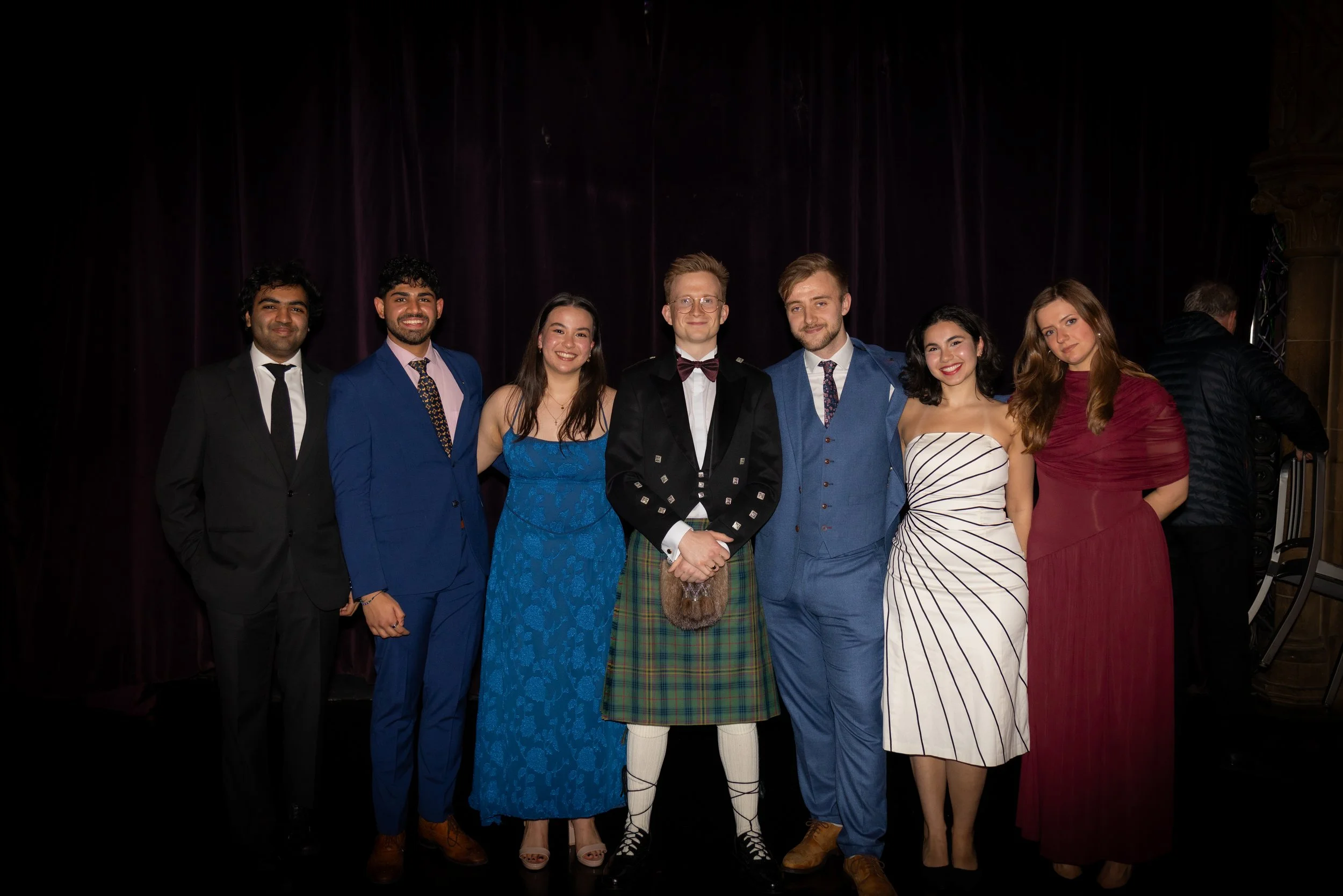 Group of eight young adults dressed in formal and traditional attire standing together on a stage with a dark curtain backdrop, smiling at the camera.
