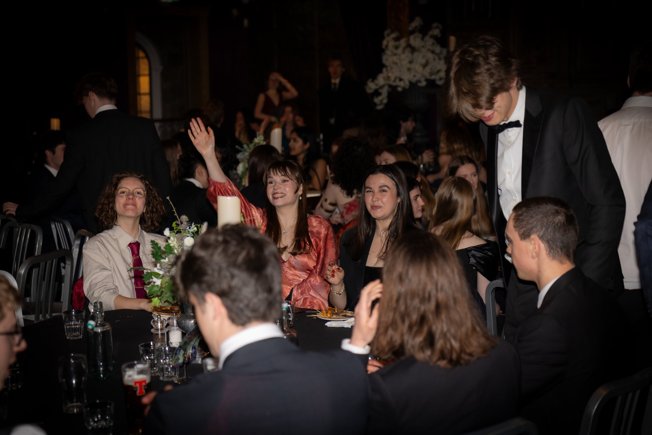 People enjoying a formal event or celebration, seated at tables with floral centerpieces, some smiling and waving, in a dimly lit room with elegant decor.